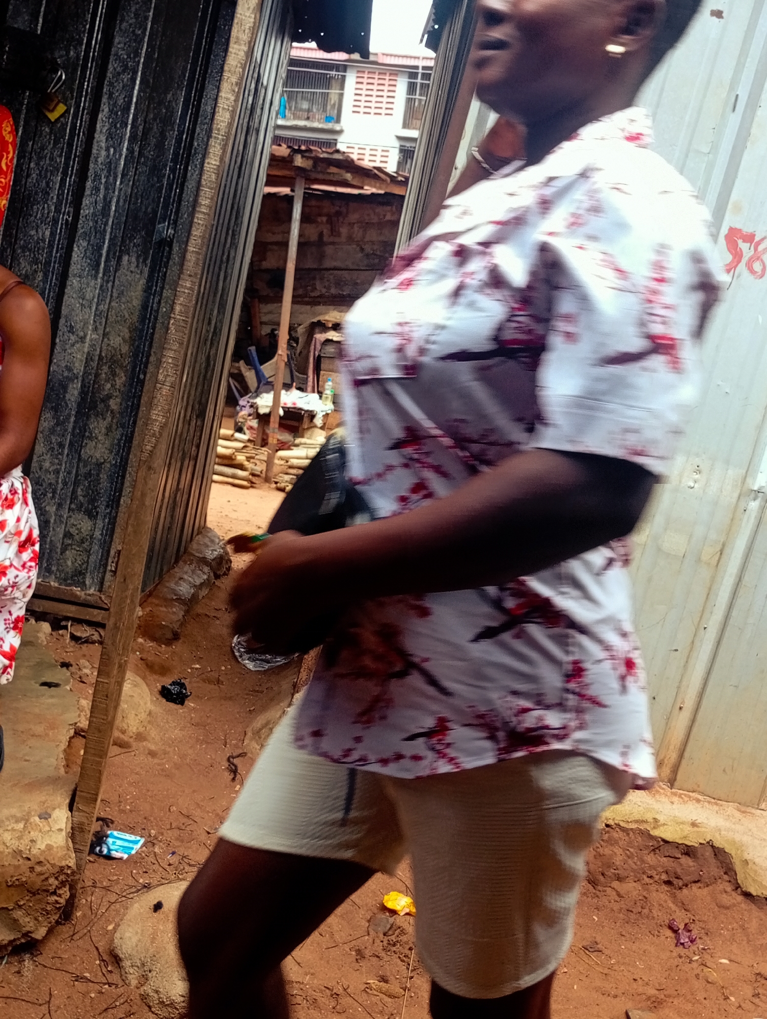 A medium shot captures a person from the chest down, standing outdoors in what appears to be a market or street environment in Nkpor, Nigeria. The individual is wearing a light-colored shirt with a red floral pattern and light-colored shorts. The background reveals a corrugated metal wall on the right and a similar structure or stall on the left, with debris and red-brown earth on the ground. The scene suggests a casual, possibly commercial setting.