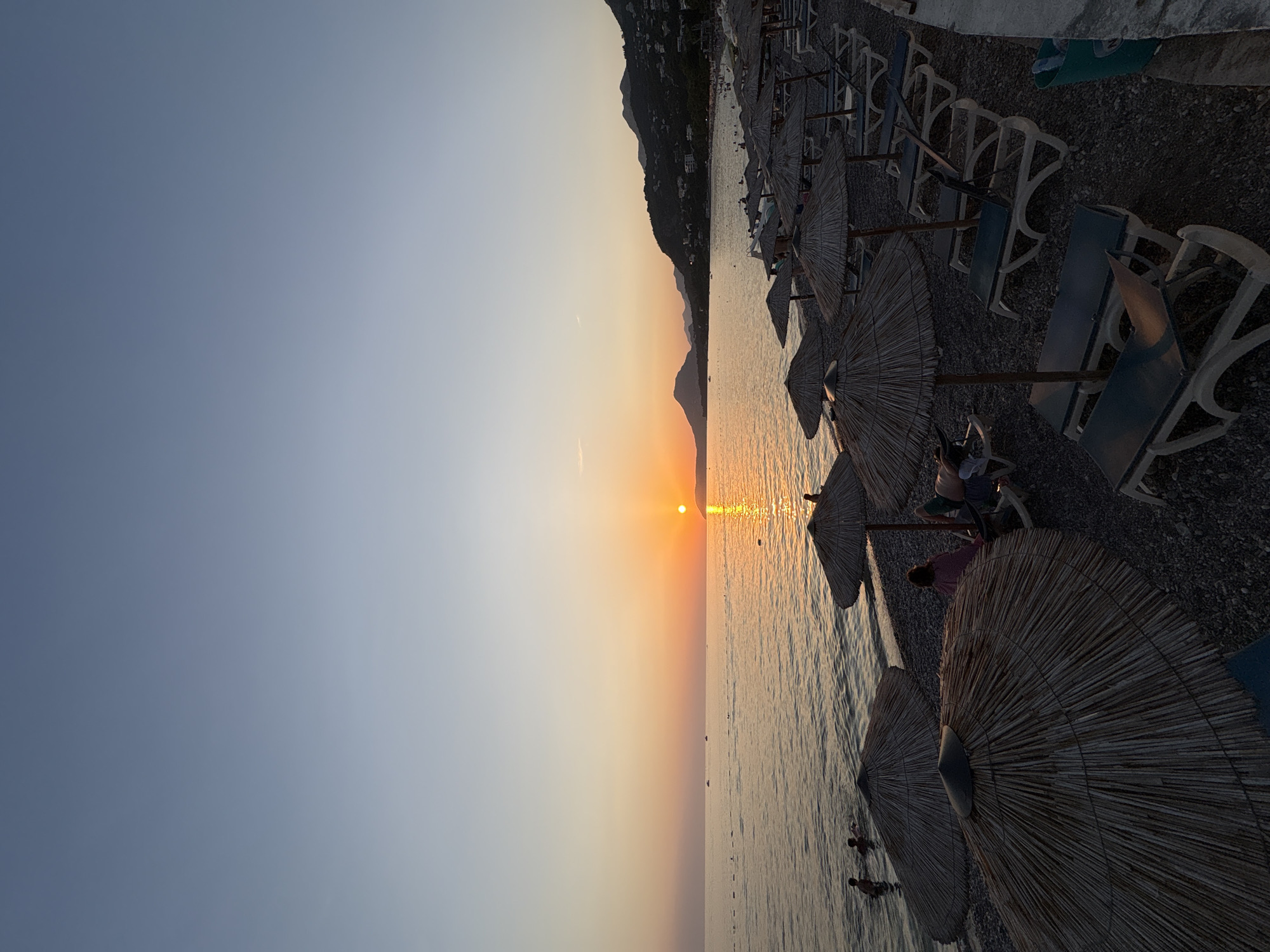 The image depicts a serene sunset scene at a pebble beach in Bar, Montenegro. The sun, a bright orange orb, is low on the horizon, casting a warm glow across the calm sea. The water reflects the golden hues of the sunset, creating a shimmering path.
On the beach, numerous thatched umbrellas and empty lounge chairs are arranged in neat rows, suggesting a popular tourist spot. A few people are visible in the shallow water, enjoying a swim. In the background, rolling hills covered with lush vegetation and dotted with houses rise up, forming a picturesque backdrop to the coastline. The sky transitions from a pale blue at the top to a soft orange and pink near the horizon, indicative of the twilight hour.