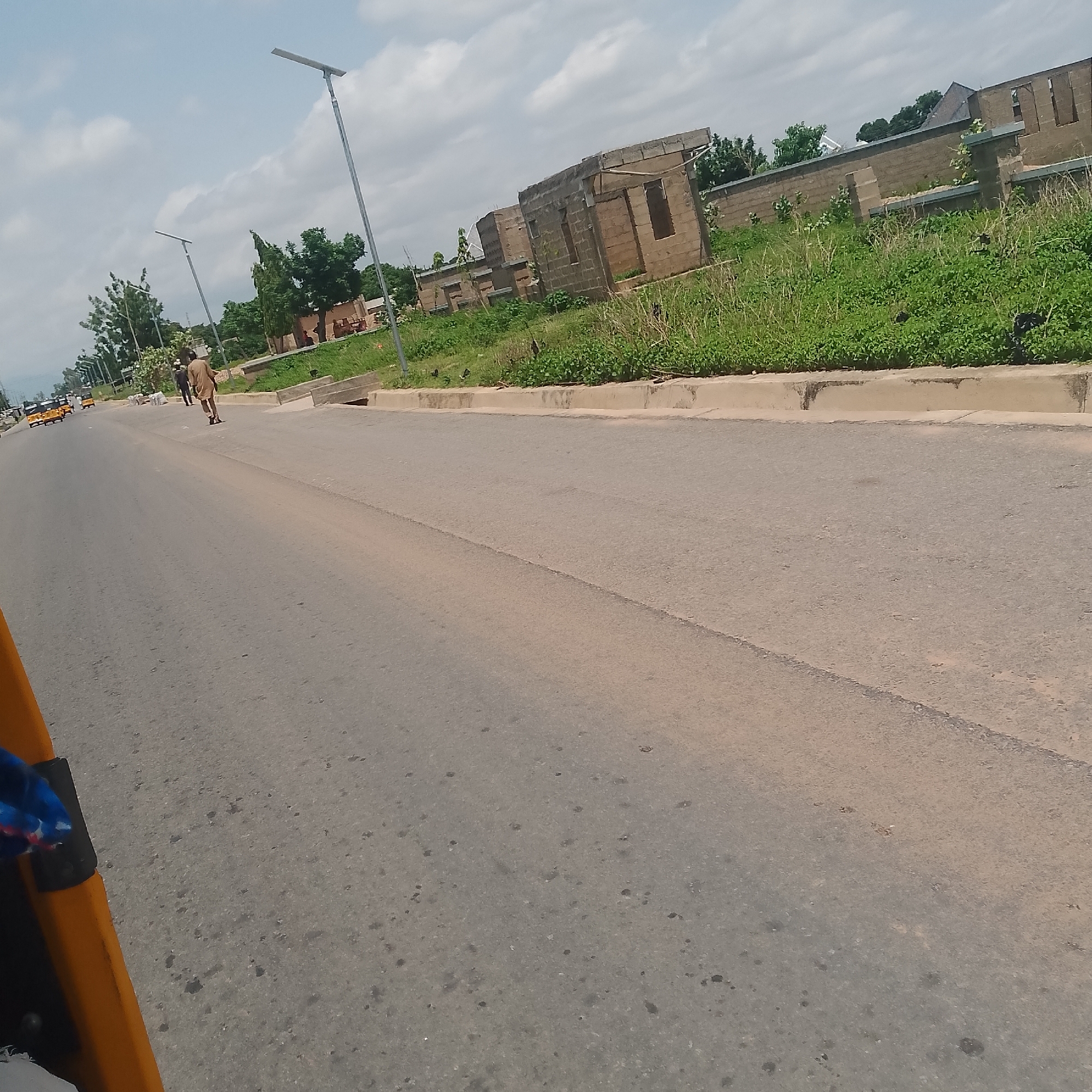 The image shows a street scene in Jalingo, Nigeria, under a partly cloudy sky. In the foreground, the paved road surface occupies a significant portion of the frame, with a faint double yellow line marking the center. To the left, the side of a yellow vehicle, possibly a tricycle or "Keke Napep," is visible, with a blue and red patterned object or fabric in the corner.
Further down the road, two individuals are walking away from the camera. The person in front is wearing a light brown traditional outfit. Behind them, another person in a darker outfit is also walking. The road is lined with a concrete curb and a grassy verge.
Beyond the verge, construction is evident. Buildings made of concrete blocks, some unfinished, are visible. The structures have exposed rebar and incomplete walls and rooflines, indicating ongoing construction. Greenery, including grass and some trees, grows around the construction site and along the roadside.
Streetlights with solar panels are installed along the side of the road. In the distance, more vehicles and a line of trees are visible, suggesting a suburban or developing area within Jalingo. The overall impression is a mix of everyday urban life and ongoing development.