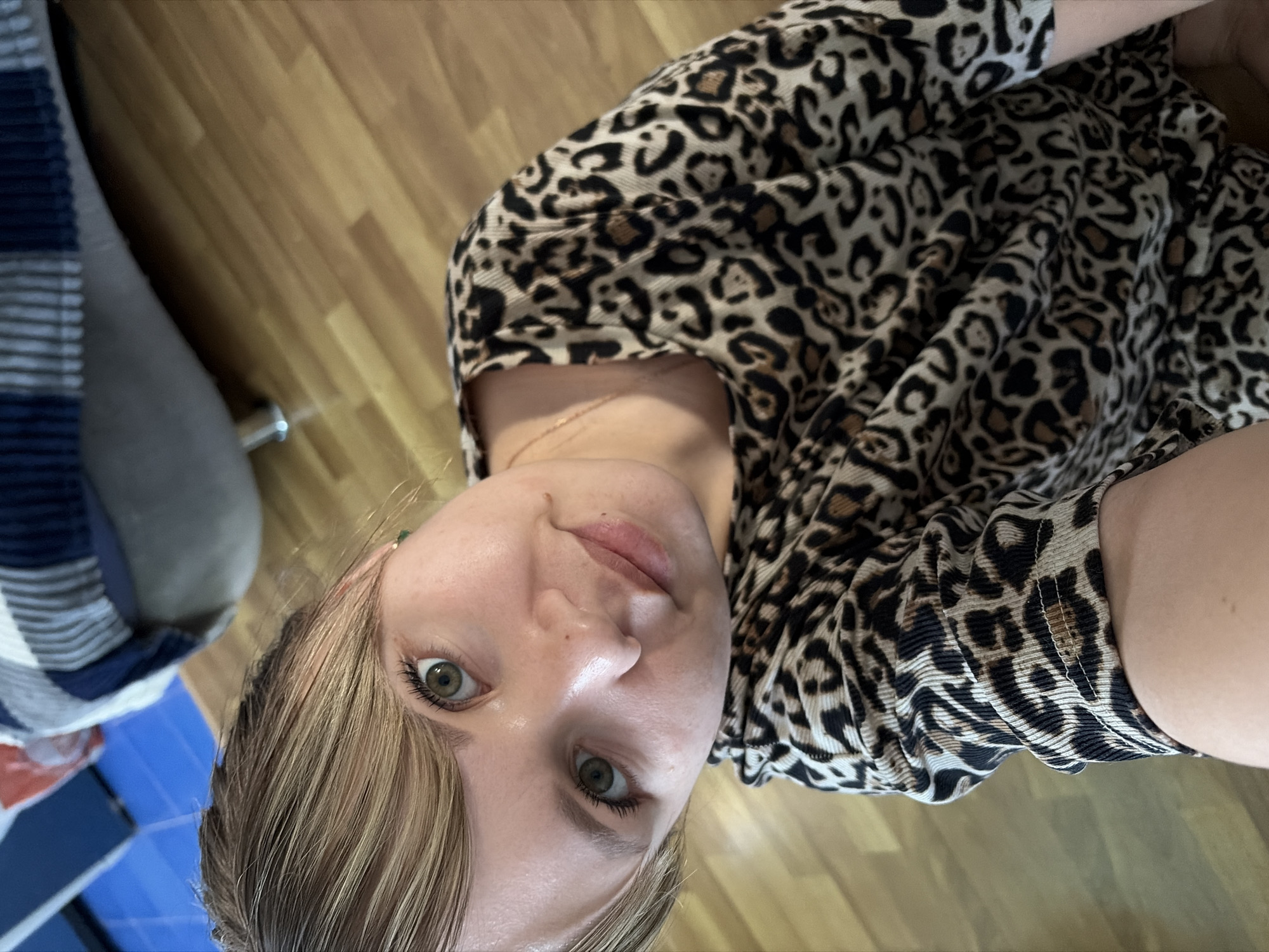 A medium-shot, close-up photograph presents a woman wearing a leopard-print top, with a possible glimpse of a furniture piece featuring blue and white stripes and a light-colored cushion positioned in the left frame. The flooring is made up of light-colored wooden planks. While the context of the location cannot be determined to be located in the city of Chaikovsky, Russia. The picture is captured from an elevated, slightly rotated perspective. The environment appears to be indoors.