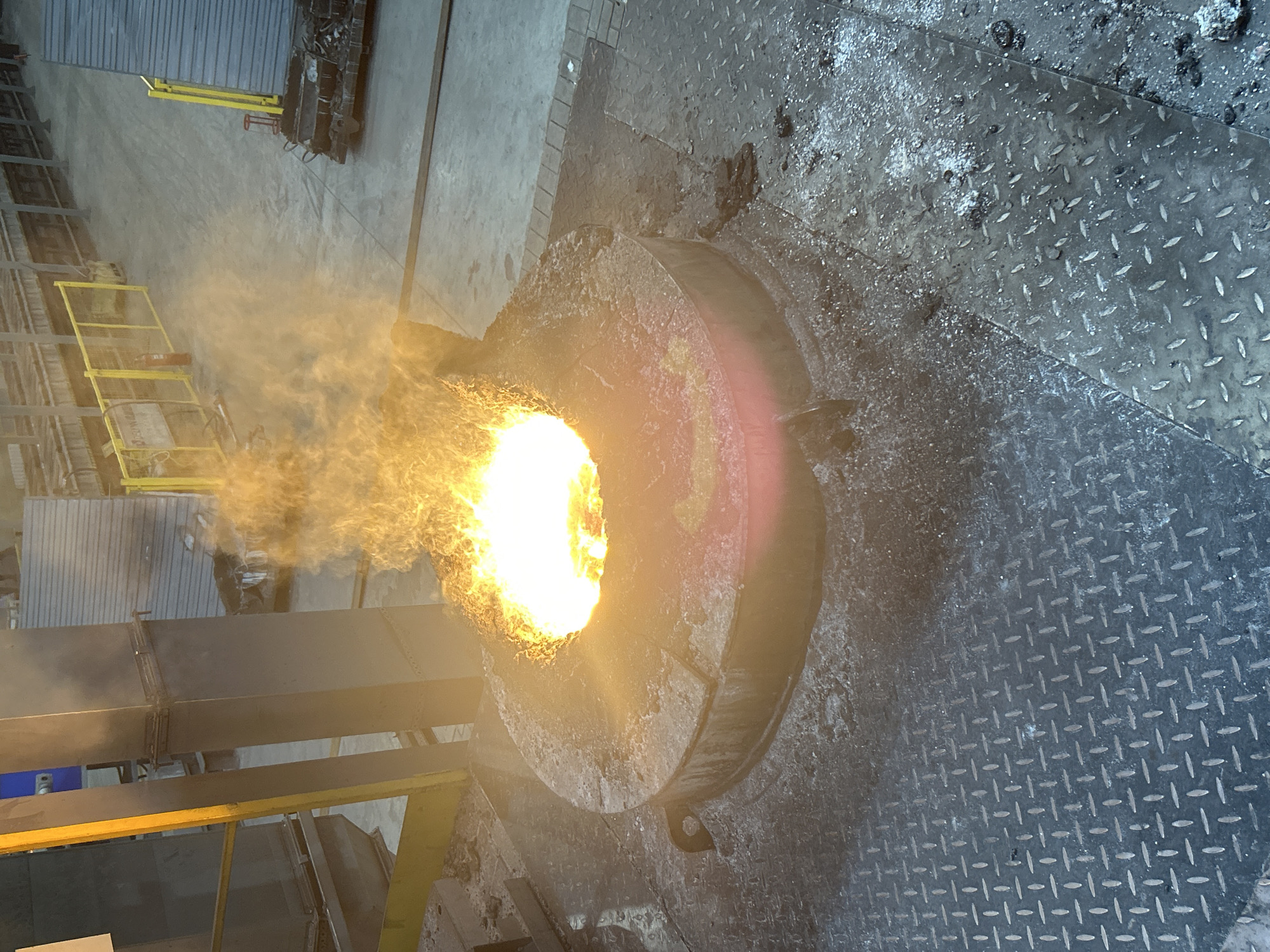 An overhead shot shows a metal foundry with a furnace containing molten metal. The furnace is surrounded by a diamond-plated steel floor. Smoke and flames rise from the furnace opening. Structural steelwork with yellow safety railings is visible to the left of the furnace. The location of Noviy, Russia is not evident from this image.