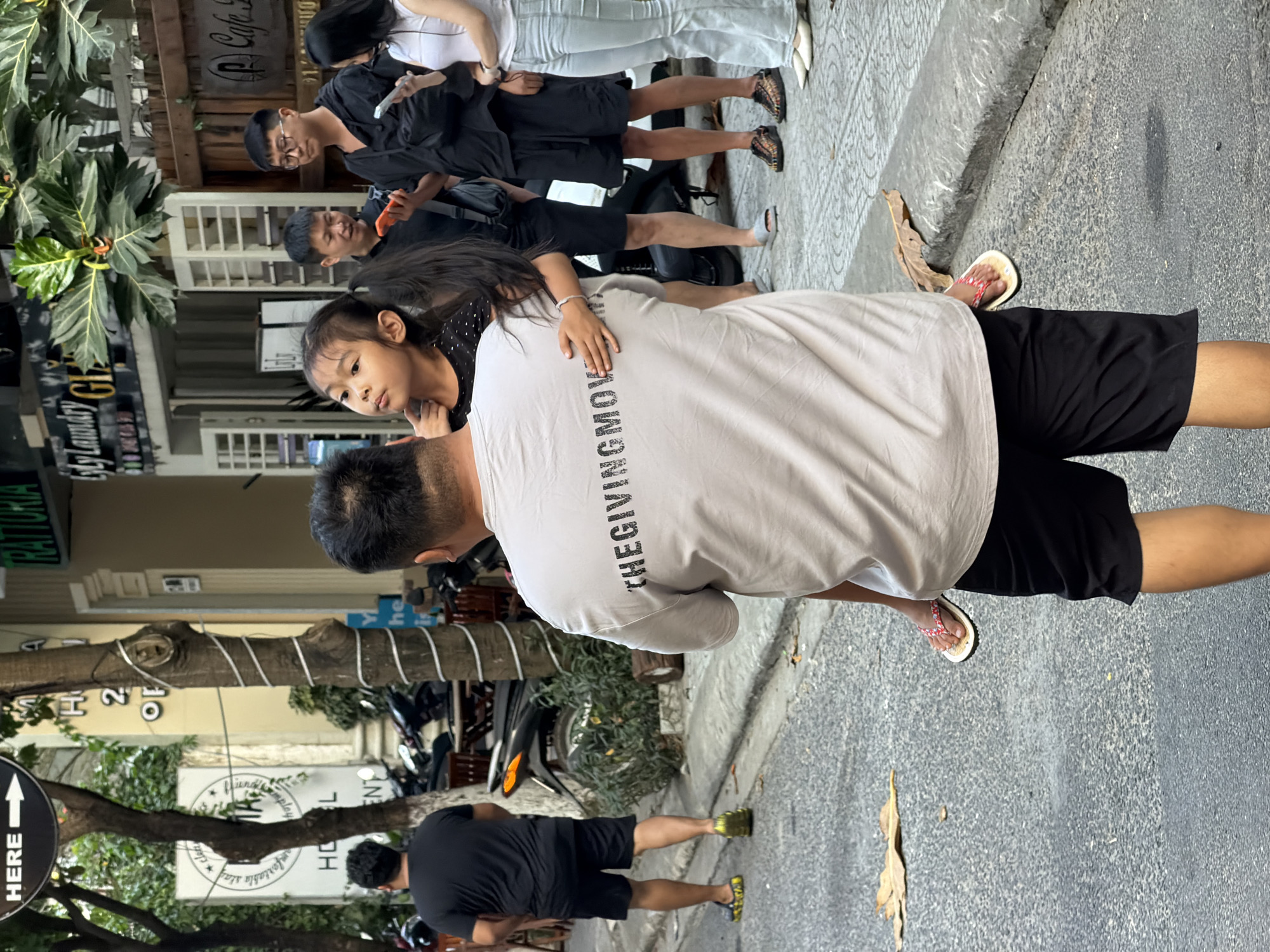 The image presents an outdoor urban scene, predominantly showing a man with a young girl in what appears to be a familial embrace. The man is wearing a light-colored t-shirt with the text "THEGIVINGMOV" printed on the back, paired with black shorts and flip-flops. The girl is dressed in a black top with white polka dots.
Several other people are visible in the background, including individuals in black clothing and one person in white pants and a white top. A street with a sidewalk forms the ground, and there is a scooter parked nearby.
Signs and storefronts are visible, including one that reads "byly Laundry Chr" and "PCafe So," suggesting a commercial area. There is also a signpost with an arrow pointing towards "HERE." A tree with lights wrapped around it is also visible.
Based on the text "RATTORIA" and the mention of the location, the image is likely taken in Đà Nẵng, Vietnam. The scene depicts a street setting with people interacting and commercial establishments.
