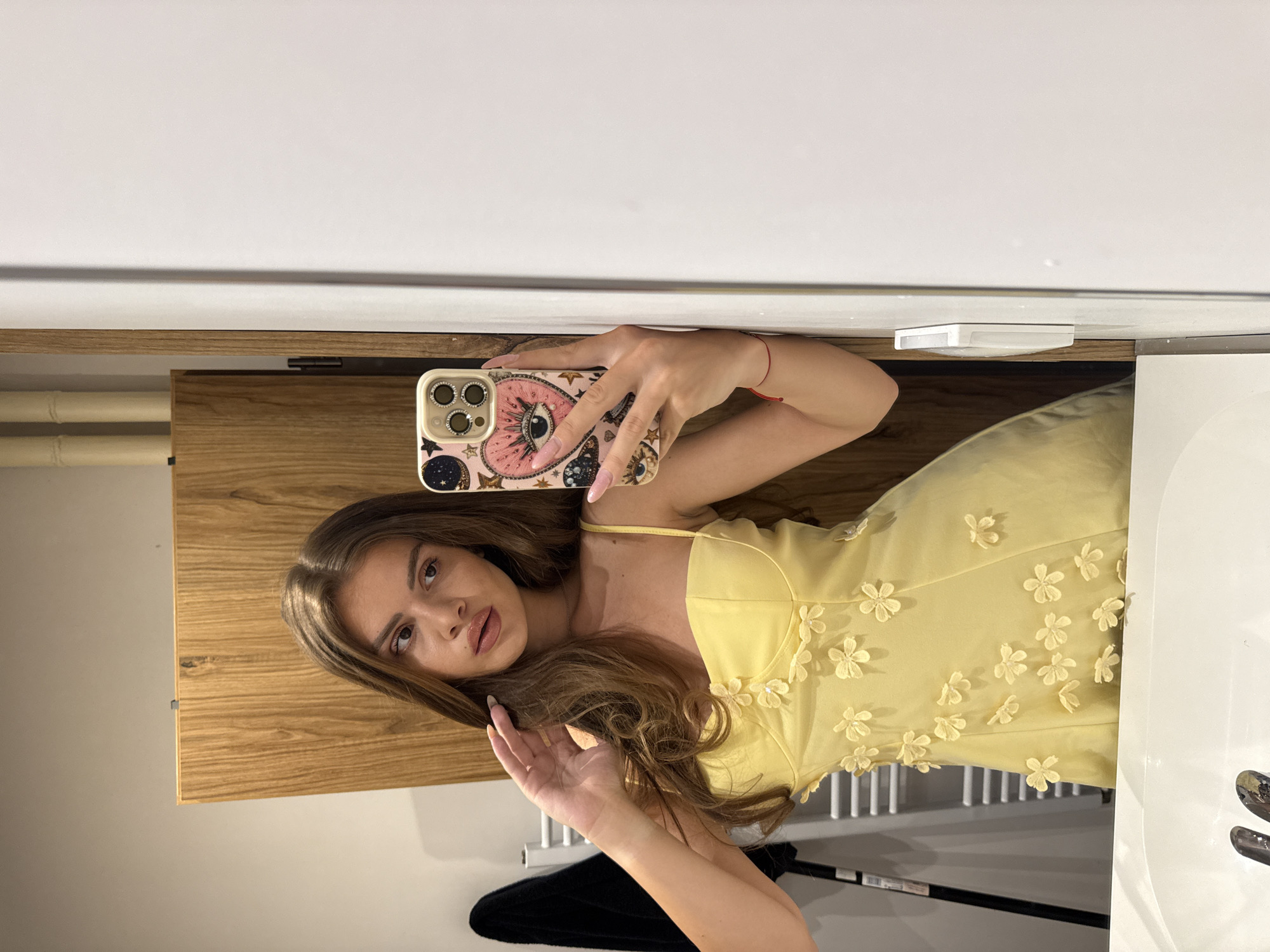A young woman with long, brown hair is taking a mirror selfie in what appears to be a bathroom. She is wearing a yellow dress adorned with small floral embellishments. The background includes a wooden cabinet, a white wall, a sink, and a towel rack. The woman is holding a phone with a decorative case. There is a red string bracelet on her left wrist. The scene does not provide any specific indicators that would place it in the city of Plovdiv, Bulgaria.
