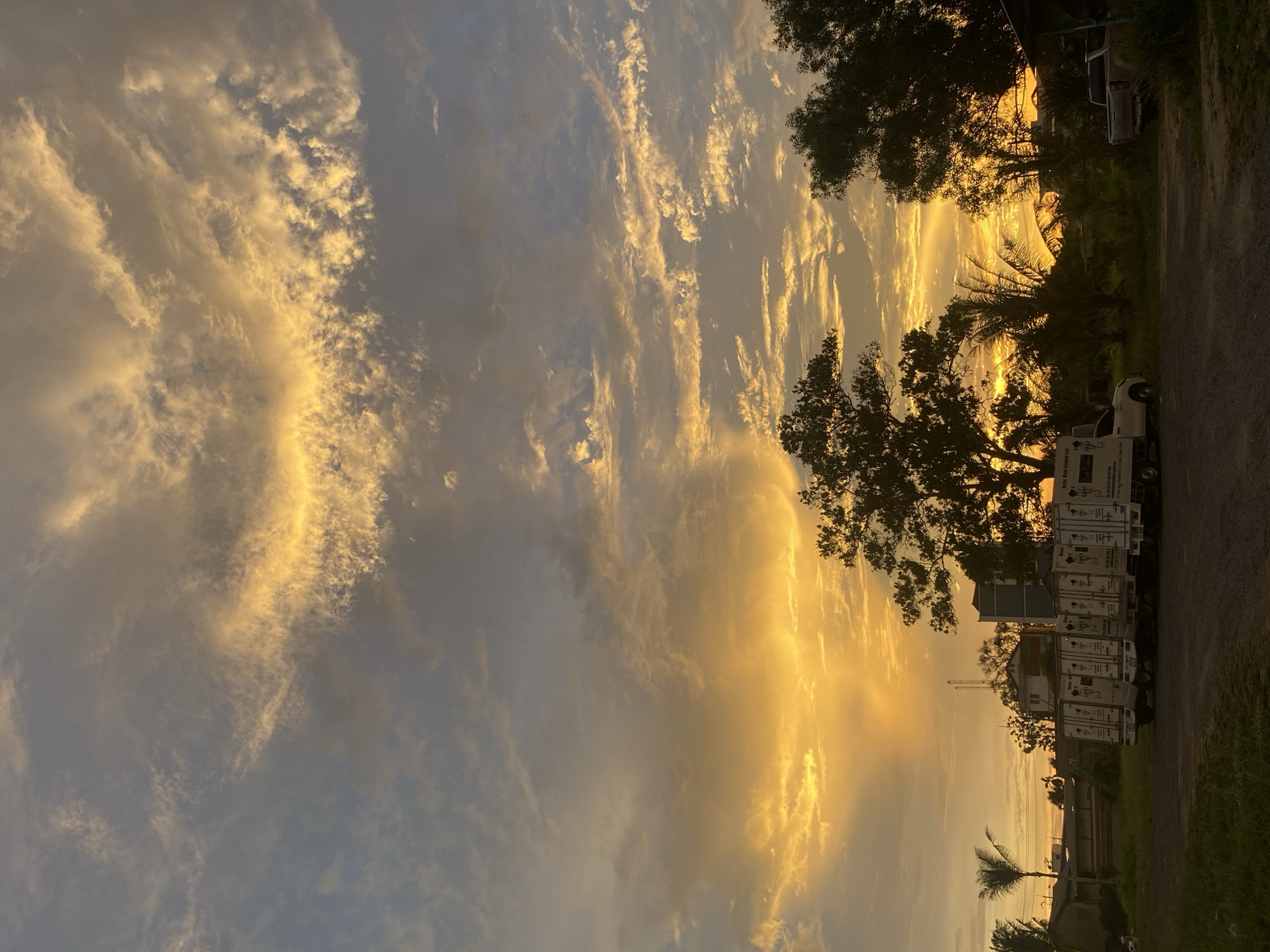 The image depicts a vibrant sunset over what appears to be the city of Choeng Thale, Thailand. The sky is filled with altocumulus clouds, illuminated by the golden light of the setting sun. A line of box trucks is parked on the side of a road, partially obscuring palm and other trees. A crane can be seen in the distance. A glimpse of buildings and residential structures is also visible. The foreground features a paved road and grassy areas, suggesting a suburban or semi-rural setting. The light is warm and intense, indicative of the tropical climate.