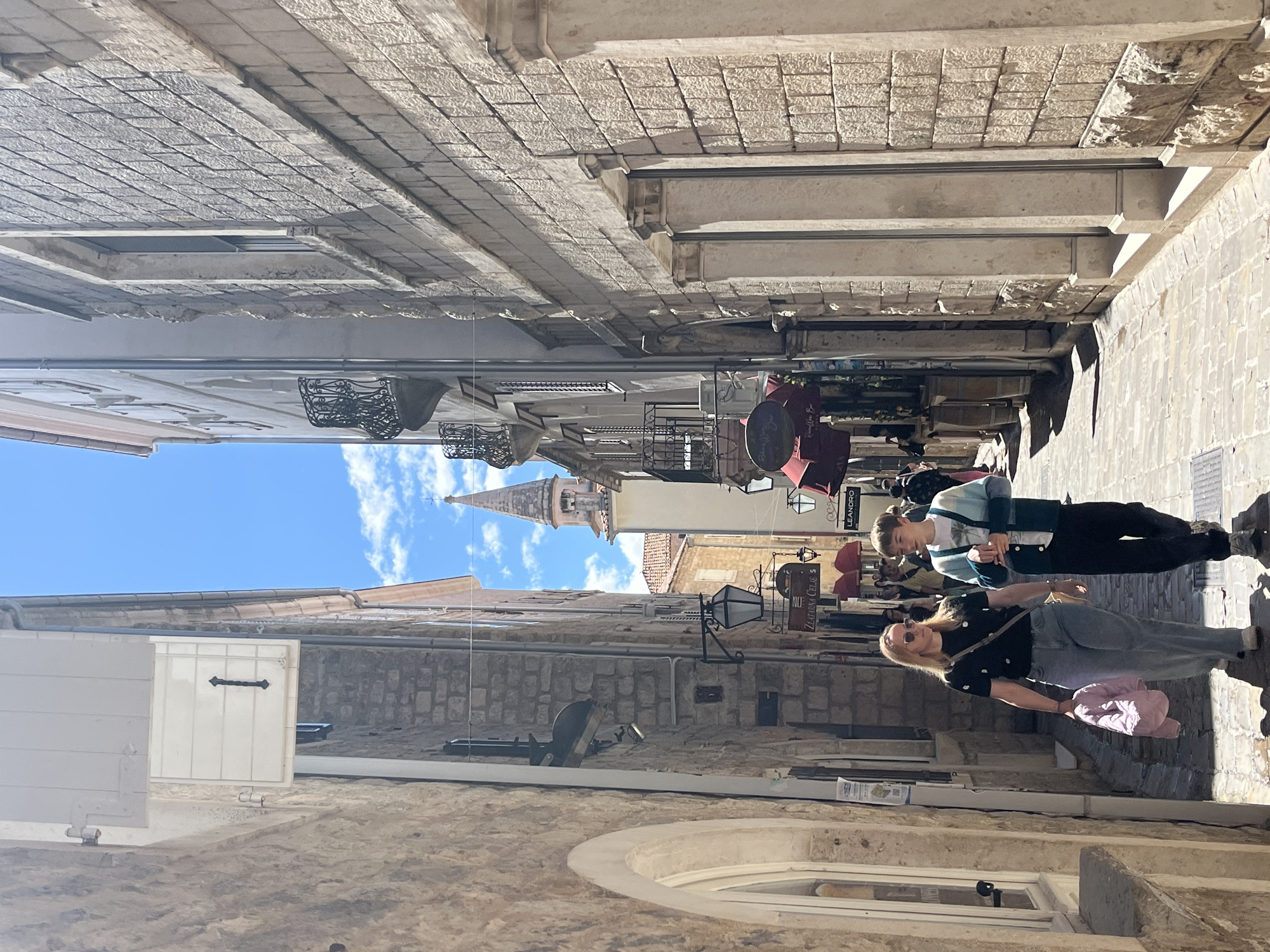 The image shows a narrow street in the old town of Budva, Montenegro. The street is paved with light-colored stone. Tall buildings with stone walls line the street on both sides. Two people are walking down the street. The person closest to the camera is a woman with blonde hair. She is wearing sunglasses, a black shirt, and light blue jeans. She is carrying a pink bag. The other person is wearing a light blue and dark blue sweater, a white shirt and dark pants. A tower can be seen in the distance between the buildings. There are signs and awnings hanging on the buildings. A window is visible in the bottom left corner of the image.