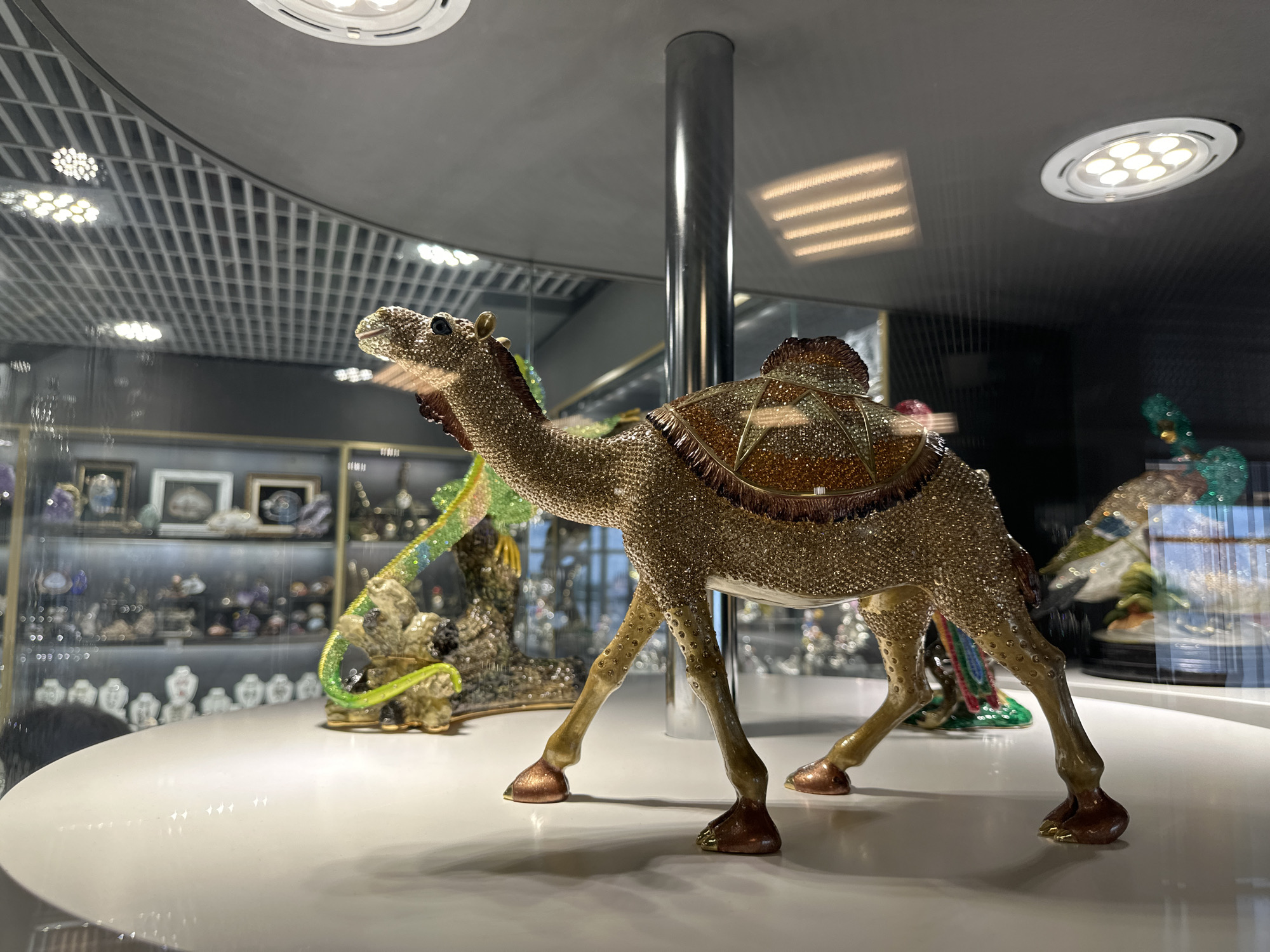 The image shows a decorative camel figurine on display in what appears to be a museum or high-end retail setting. The camel is adorned with a mosaic of sparkling crystals or similar materials, giving it a luxurious appearance. It has a saddle on its back, also elaborately decorated.
The figurine is placed on a white, circular display table. The background reveals shelves filled with other decorative objects and possibly jewelry, suggesting the location is a store or museum exhibit. The lighting is bright, reflecting off the polished surfaces of the display. The room features a modern ceiling with a grid-like structure and recessed lighting.
Given the subject matter and style, the location could potentially be within a retail or museum space in a city like Saint Petersburg, Russia, known for its rich history and art. However, without further visual clues, it's not possible to confirm the exact location with certainty.