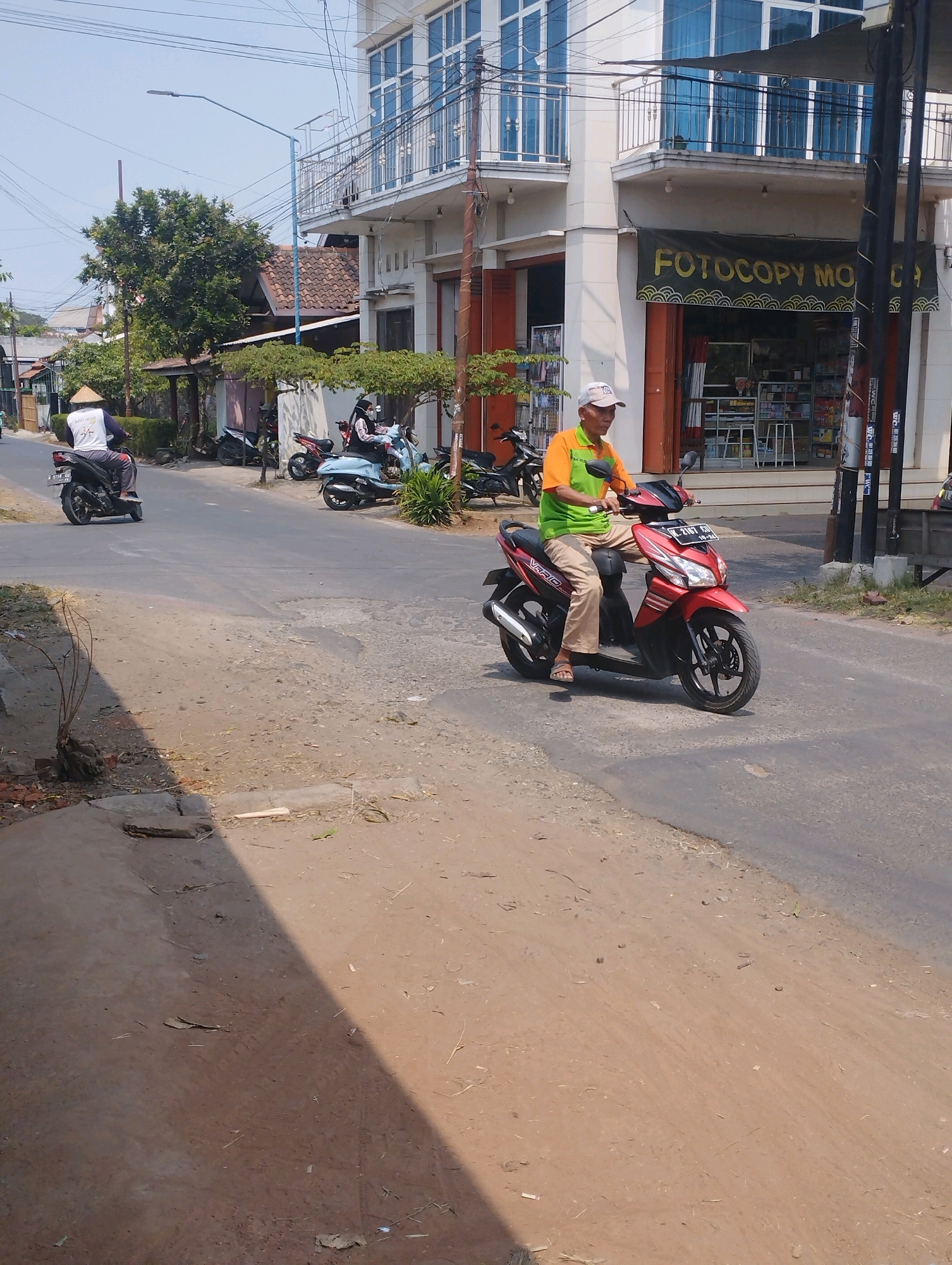 The image depicts a street scene in Trangkil, Indonesia, on a sunny day. In the foreground, a man wearing a light-colored cap, a green and orange t-shirt, and tan pants is riding a red scooter. He is looking forward. The scooter has a license plate with the characters "L 2167 CH". Behind him, on the left side of the road, a person wearing a white shirt and a conical hat is riding a black scooter. Further down the street, other scooters are parked and in motion, including a light blue scooter with two people on it, one wearing a face mask.
The buildings lining the street are multi-story structures. One building prominently features a banner with the text "FOTOCOPY MO...". The street surface is a mix of asphalt and dirt. Trees and vegetation are visible along the roadside and between buildings. The sky is clear and blue. The overall impression is that of a typical urban or semi-urban environment in Indonesia.