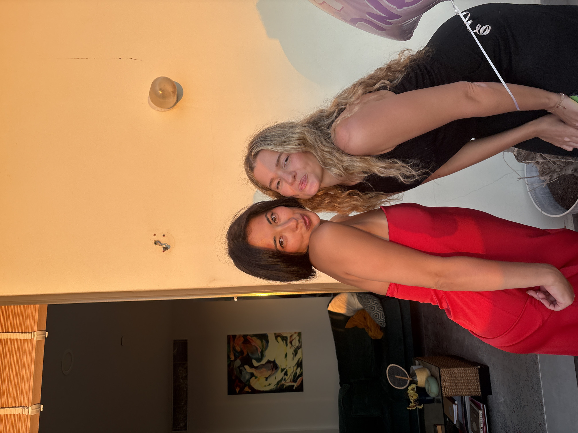Two women pose for a photo against a beige wall. The woman on the left wears a red strapless dress and pouts her lips. The woman on the right has long blonde, curly hair and wears a black sleeveless dress, making a kissing face. A portion of a purple balloon is visible on the right side of the frame, tethered by a white ribbon. Behind the women, to the left, is an open doorway revealing a living room with a dark green sofa and a painting on the wall. The scene appears to be an outdoor setting, possibly a balcony or patio, in Tel-Aviv, Israel, given the lighting which suggests late afternoon or early evening sun. The overall ambiance is casual and celebratory.