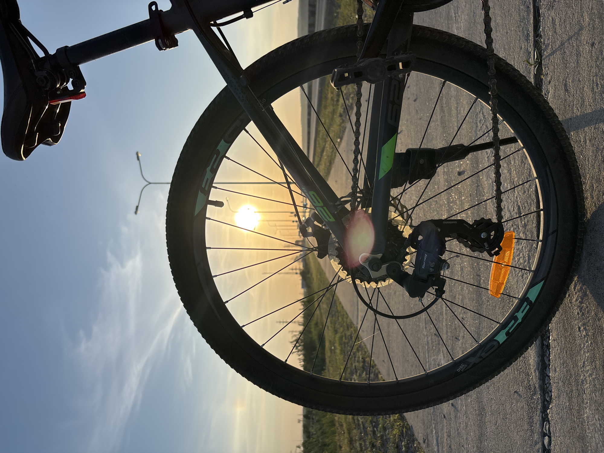 A bicycle is positioned on a concrete surface, likely a sidewalk or road, with the setting sun visible between the spokes of the front wheel. The sky is a pale blue transitioning to a yellow-orange hue near the horizon, indicating either sunrise or sunset. A distant street lamp is visible in the background. The bicycle is black with some green accents, and it appears to be a mountain bike or similar style. A chain is hanging down alongside the wheel. An orange reflector is attached to the wheel, close to the rear derailleur. The scene lacks indications to establish the location as Chaikovsky, Russia.