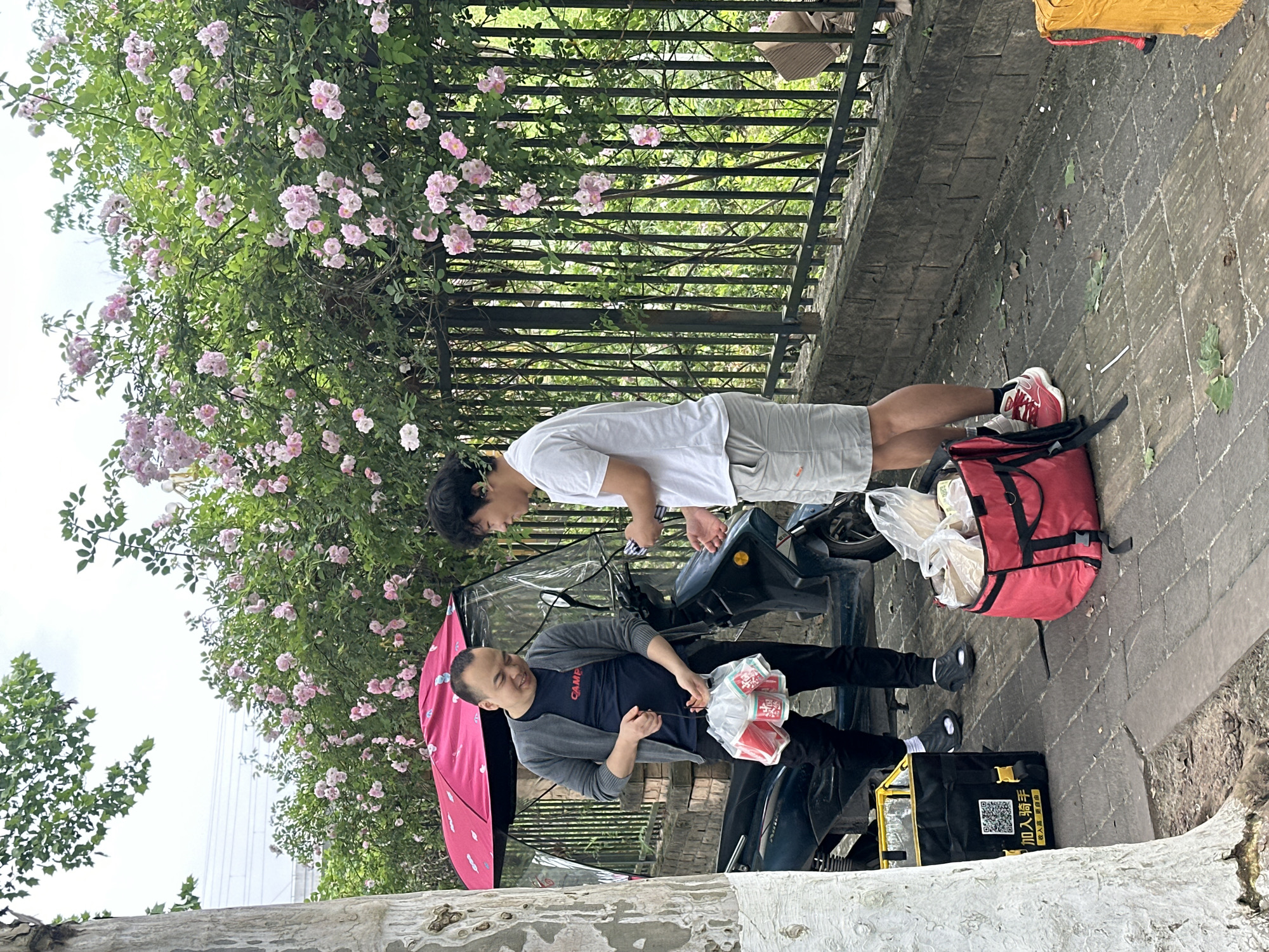 Two Asian men stand next to a black three-wheeled motorcycle on a paved path. The man in the foreground wears a gray cardigan over a dark shirt and black pants, and holds packages of food. The man in the background wears a white shirt and gray shorts, and leans forward, appearing to interact with the motorcycle. A pink umbrella is attached to the motorcycle. A red delivery bag and a black and yellow delivery bag sit on the path nearby. Flowers grow on the other side of a black fence behind them. The motorcycle and bags suggest a food delivery service. The scene appears to be located in Jiangjin, China.