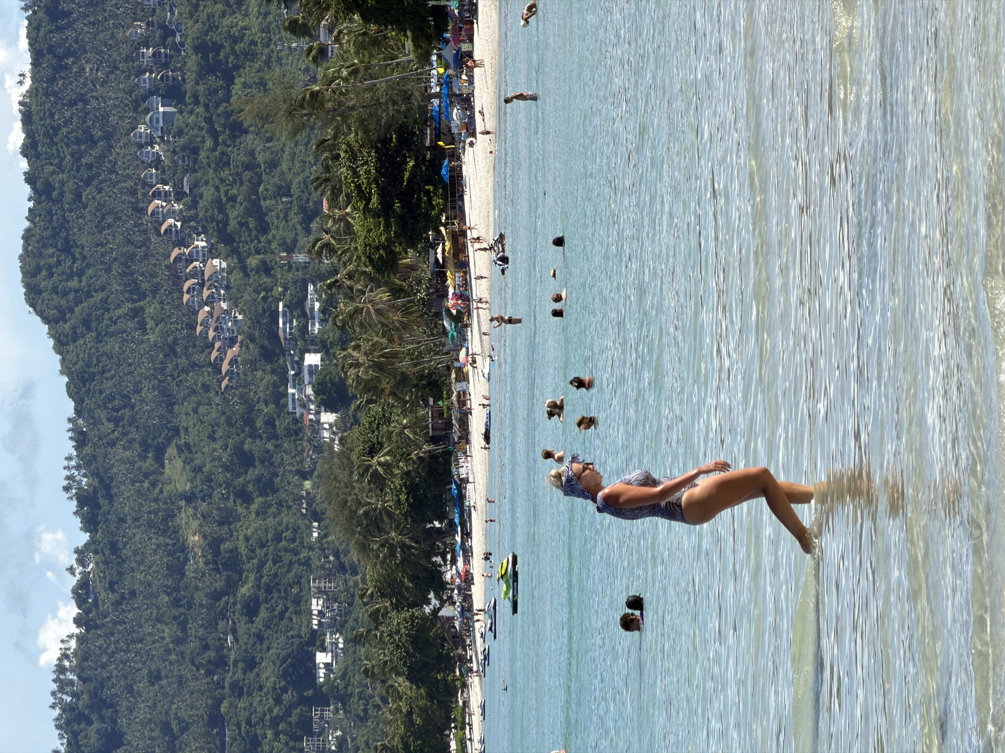 This is a vertical, high-angle shot of a tropical beach in Baan Chaweng, Thailand. The image captures a vibrant scene with a clear division between the lush green, densely forested hillside and the calm, shallow, blue water.
On the hillside, numerous white buildings, likely resorts or villas, are nestled among the trees, creating a striking contrast with the natural landscape. Palm trees are abundant, dotting the hillside and lining the beach.
The beach itself is bustling with activity. People are seen scattered across the sand and in the water. Several individuals are wading or swimming in the clear turquoise sea. A woman in a patterned swimsuit is floating on her back in the water, with her legs slightly raised, appearing relaxed. Other people are visible in the water and on the beach, some with umbrellas providing shade. There are also several jet skis parked on the sand, indicating recreational activities. The presence of umbrellas and beach shelters suggests a sunny day, likely late morning or early afternoon, given the bright lighting and distinct shadows.
The overall atmosphere is one of a leisurely tropical vacation. The water is calm and inviting, and the scenery is picturesque, characteristic of a popular tourist destination. No visible text is present in the image.