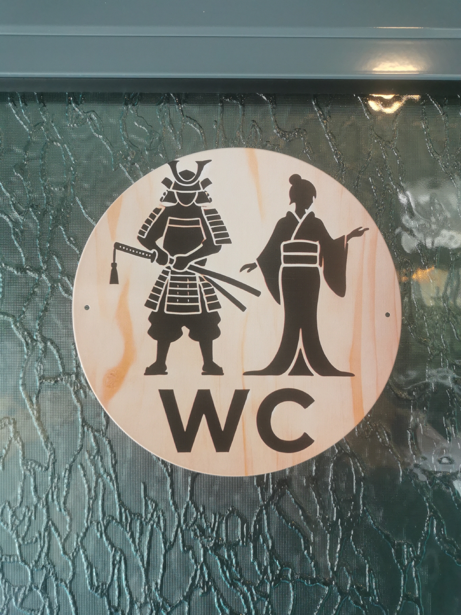 A circular wooden restroom sign, prominently featuring black silhouette figures, is mounted on a textured, translucent glass panel, with a teal-colored frame visible above. The scene is located in Blagoevgrad, Bulgaria.
The sign is made of light-colored wood with a visible grain, and it displays two stylized figures. On the left, a male figure is depicted as a samurai, wearing traditional armor, including a distinctive helmet with crescent horns (kuwagata), and carrying two swords (katana and wakizashi) at his waist. On the right, a female figure is shown gracefully in a traditional Japanese kimono with wide sleeves and a hair bun, with her left hand slightly raised. Below these cultural figures, the letters "WC" are centrally placed in large, black, sans-serif capital letters, indicating a Water Closet or public restroom.
The sign is attached to the glass panel, possibly with small fasteners visible as dark dots near the edges of the circle. The glass itself has a pronounced, irregular, almost cracked or frozen-like texture, which obscures visibility while allowing diffused light to pass through. This unique design, combining Japanese cultural motifs with a universal restroom symbol, suggests a themed establishment. No specific activity is taking place, as it is a static sign providing direction. The time of day and weather are not discernible, but a subtle reflection is visible on the teal frame above the glass.