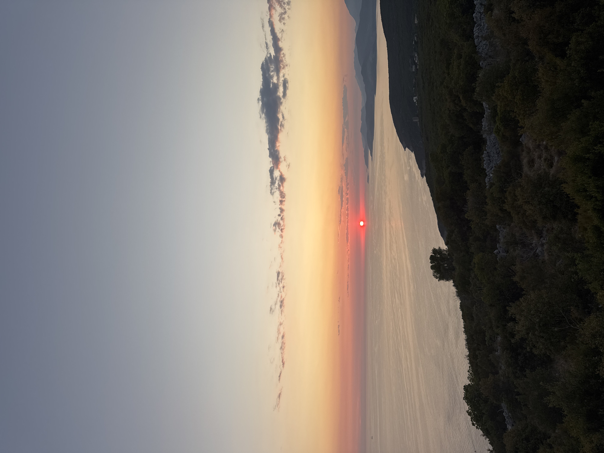 This image captures a sunset over the Adriatic Sea, as viewed from a hillside overlooking the city of Kovačko Polje, Montenegro. The sun, a vivid red orb, is setting on the left side of the frame, casting a warm glow across the water and the sky. Wisps of pink and orange clouds streak across the horizon. The foreground is dominated by a dense, dark green hillside covered in trees and shrubs, with a stone wall visible in the lower right portion. In the distance, the coastline curves, with faint outlines of mountains visible against the twilight sky. A few distant boats dot the water. The overall scene is serene and picturesque, typical of a Mediterranean coastal landscape at dusk.