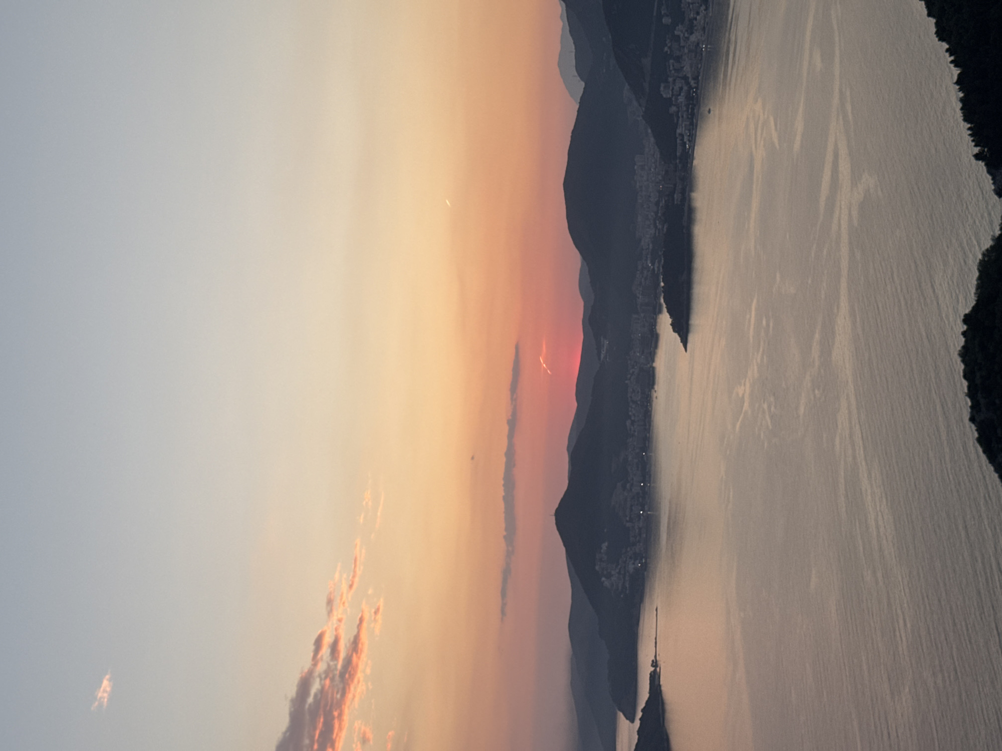 The image captures a sunset view from Sveti Stefan, Montenegro. The sky transitions from pale blue to orange, with streaks of pink clouds near the horizon. Silhouetted mountains and coastal structures are visible. The water reflects the sky's colors.