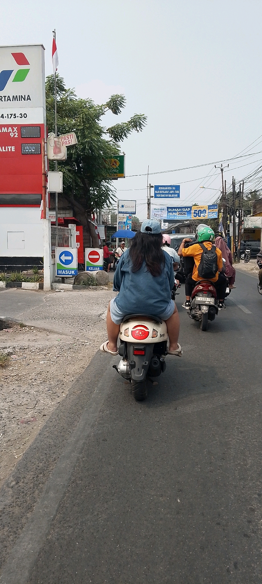In Karangsatria, Indonesia, a street scene unfolds with several motorcycles in motion. A woman wearing a denim jacket and a blue baseball cap is the central figure, riding on the back of a cream-colored scooter. She is dressed in shorts and white sandals. The scooter is following another motorcycle carrying two individuals, one wearing a green helmet and an orange shirt, and the other a brown headscarf. Further ahead, more motorcycles are visible. To the left of the frame, a red Pertamina gas station sign is prominent, displaying fuel prices and brand logos. Next to the gas station, there are traffic signs indicating "MASUK" (entrance) with a left arrow and "KELUAR" (exit) with a red circle and a horizontal bar. The environment is an urban street with asphalt paving, a dry, sandy shoulder, and utility poles with numerous wires overhead. Trees and various advertisements are also present along the roadside. The sky is overcast, suggesting a cloudy day.