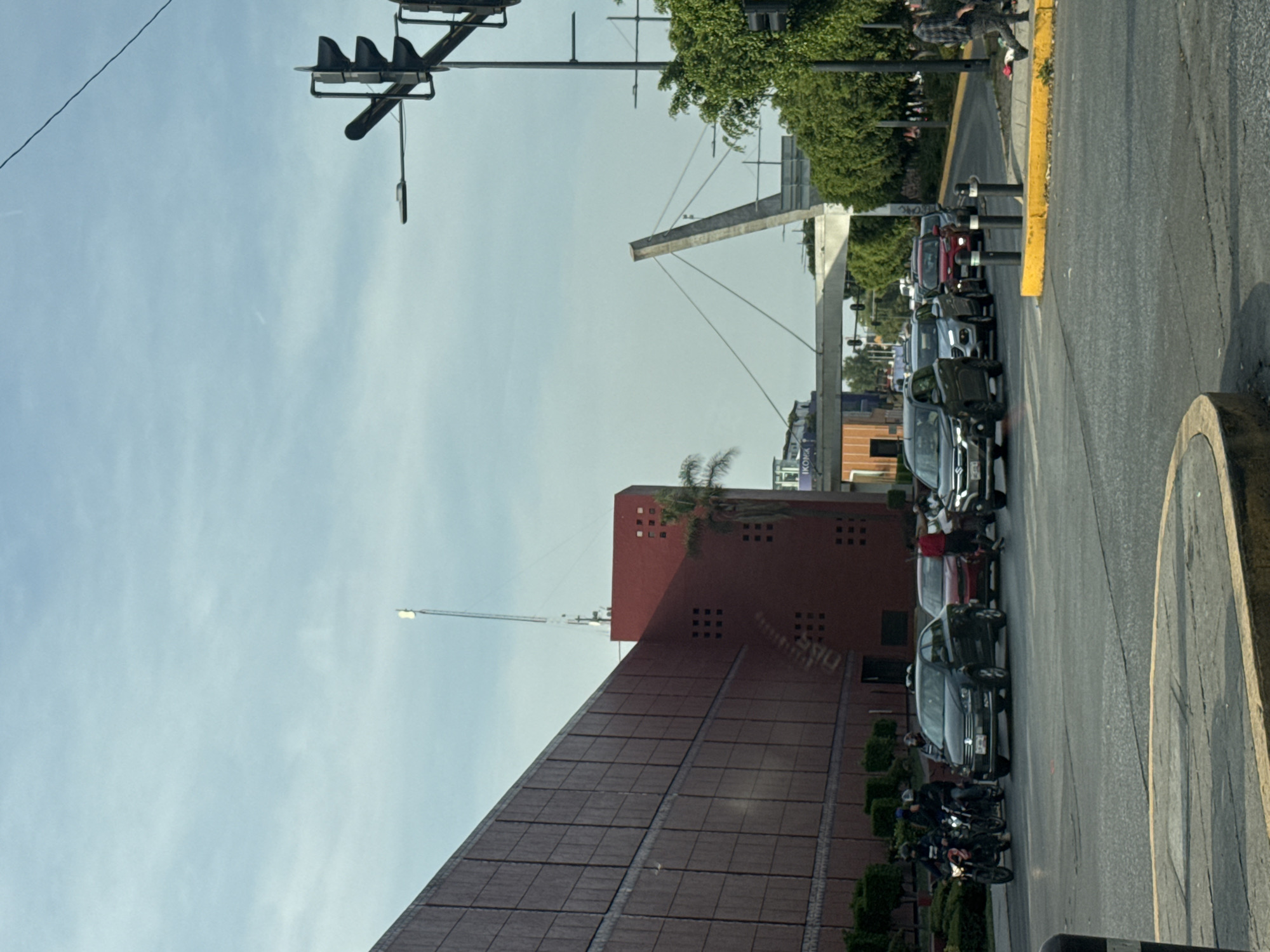 This media file captures an urban street scene in Puebla, Mexico, from the perspective of a vehicle. The image is set during daytime under a clear to lightly clouded blue sky.
In the foreground and midground, a multi-lane asphalt road stretches out, populated by a line of cars and several motorcycles, indicating either stopped or slow-moving traffic. A distinct yellow curb or median separates the road from a paved area on the right. Black and white traffic barriers are also visible along the roadside.
To the left, a prominent building with a reddish-brown, tiled facade and a steep, sloping roof dominates the scene. A more rectangular section of this building features several square windows. A palm tree grows on top of this structure, and a tall pole with a light fixture extends high into the sky.
In the background, a modern, light-colored concrete structure, potentially part of a bridge or overpass, is visible, supported by cables. Lush green trees are scattered throughout the landscape, bordering the road and buildings. Overhead, a black traffic light system is suspended.
A few people are present, including individuals riding motorcycles and some smaller, indistinct figures on a raised area in the distance. Partially visible text "IKOKA" can be seen on a sign further back in the scene. The overall impression is a busy city thoroughfare on a bright day.