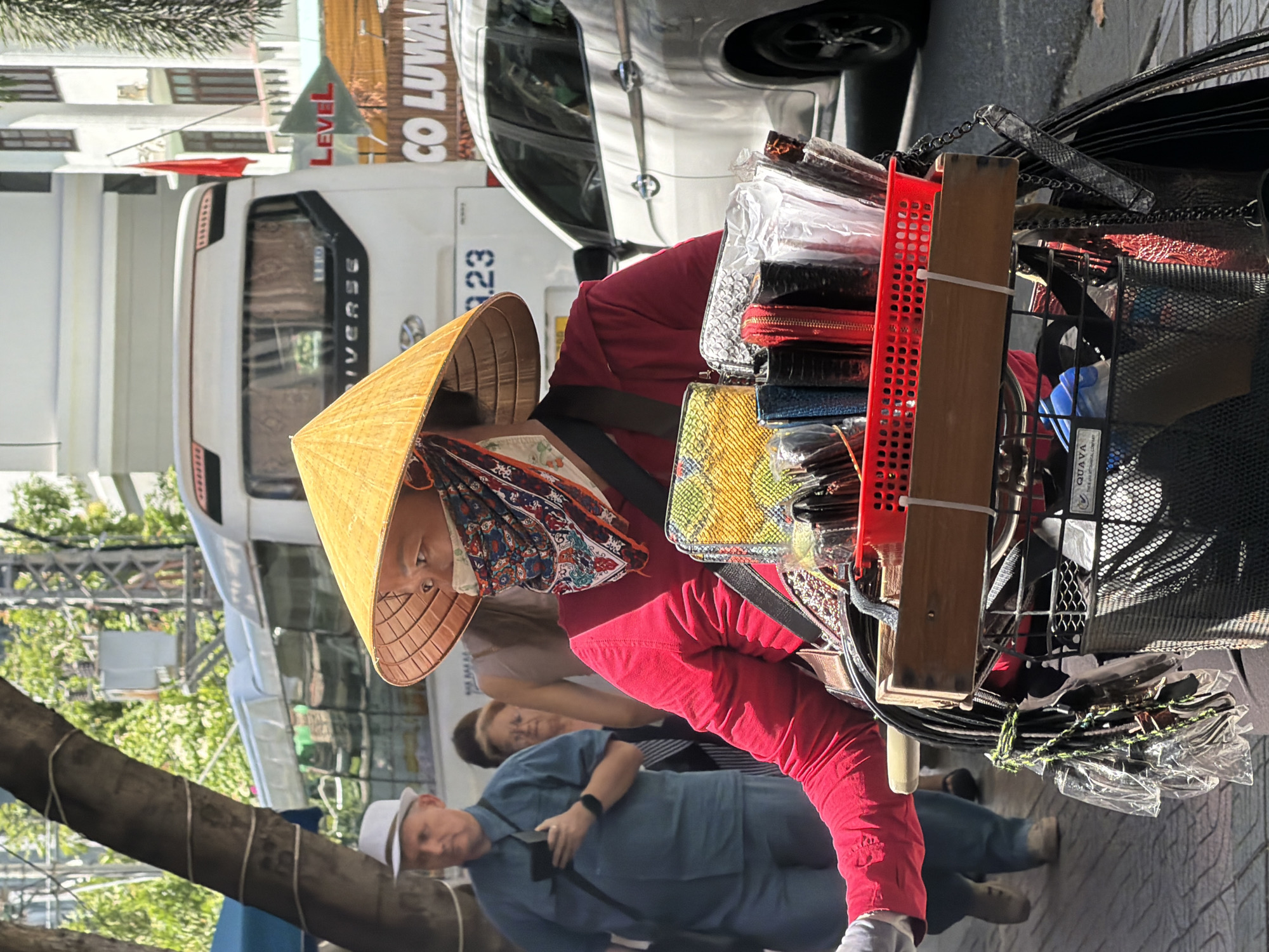 A woman wearing a conical hat and a patterned face covering pushes a cart of goods through a street in Nha Trang, Vietnam. She is dressed in a red long-sleeved shirt and has a colorful scarf covering her face. The cart is laden with various items, including purses and small bags, arranged in a red plastic basket and a wooden crate. A man in a blue shirt and white hat is visible in the background, looking at his phone. A white bus and a white car are also present in the scene, along with buildings and trees. The ground is paved with stone tiles, and sunlight suggests it is daytime. The overall scene depicts a bustling street environment.