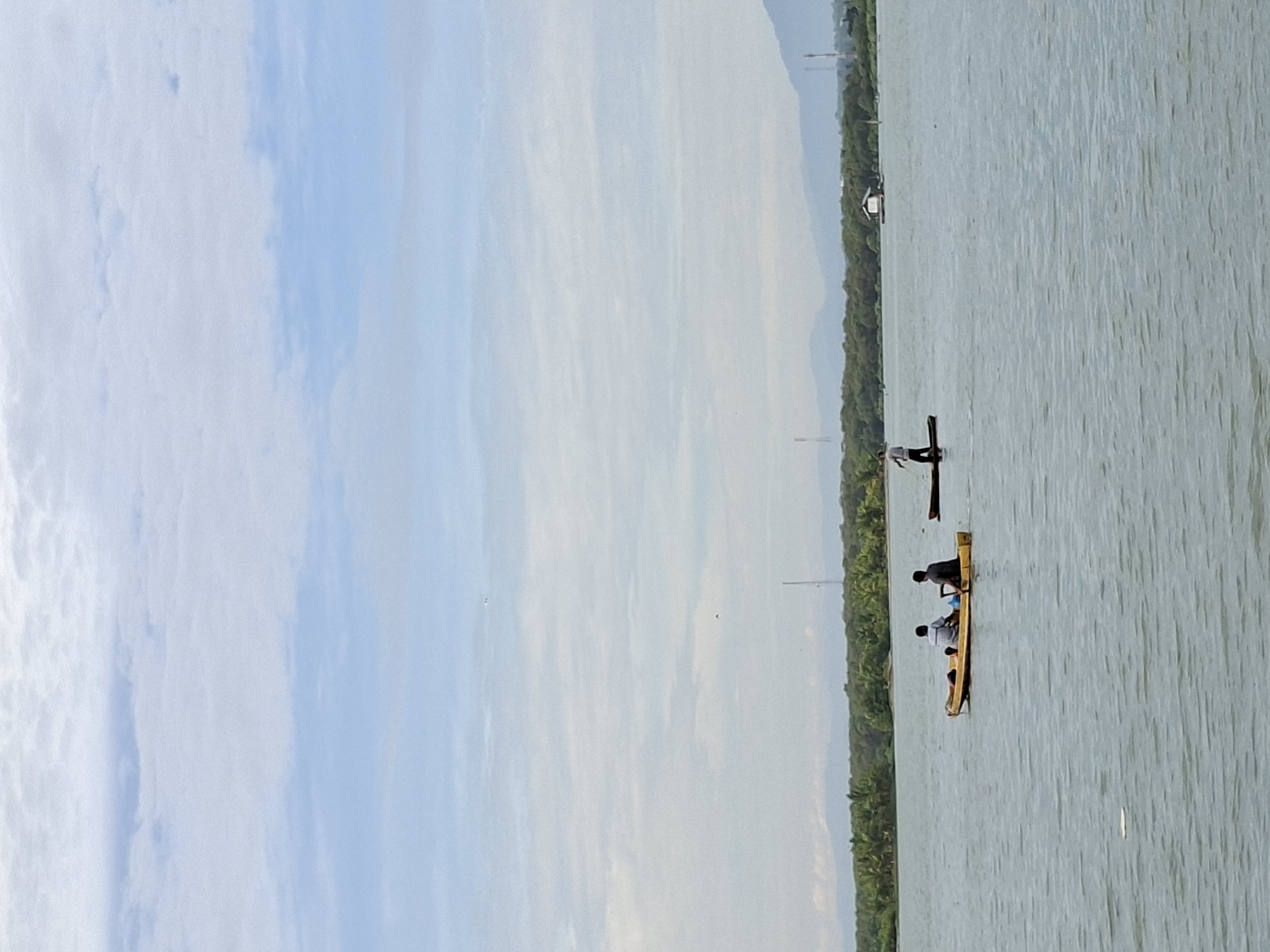 Two people are in a yellow kayak on a body of water. One person is seated in the front, and the other is seated in the back, paddling. Further in the distance, another person is standing on a paddleboard, also on the water. The background features a lush green tree line along the shore, with mountains visible in the far distance. A few structures, possibly buildings or rafts, are scattered along the shoreline. The sky is partly cloudy with a hazy appearance. This scene is likely located in Lhokseumawe, Indonesia, known for its coastal areas and natural landscapes.