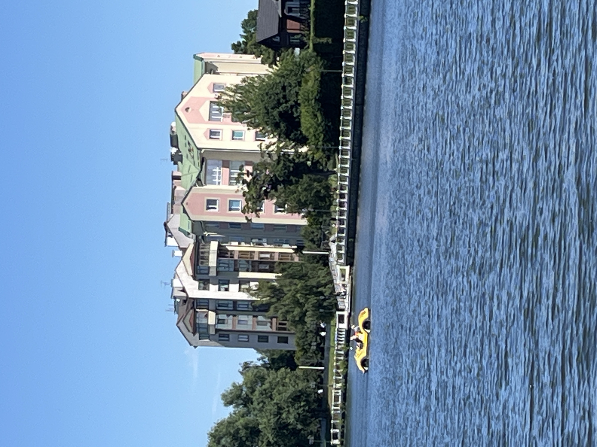 The image captures a serene waterside scene with modern apartment buildings bordering a calm body of water. The buildings are colorful, featuring pastel pink, beige, and green facades, with multiple stories and numerous windows. Lush green trees are interspersed between the buildings and along the water's edge, creating a pleasant contrast with the urban architecture. A white fence runs along the perimeter of the buildings, separating them from the water.
In the water, a bright yellow pedal boat is visible, with at least two people aboard. They appear to be enjoying a leisurely ride on the water. The water itself has a textured surface, indicating a gentle breeze or ripples.
The sky is a clear, bright blue, suggesting a sunny day, likely in the late morning or early afternoon. The overall atmosphere is tranquil and idyllic, hinting at a residential area or a recreational spot. The presence of the pedal boat and the clear weather suggest it is a pleasant time for outdoor activity. No visible text is present in the image. The context provided places this scene in Kaliningrad, Russia.