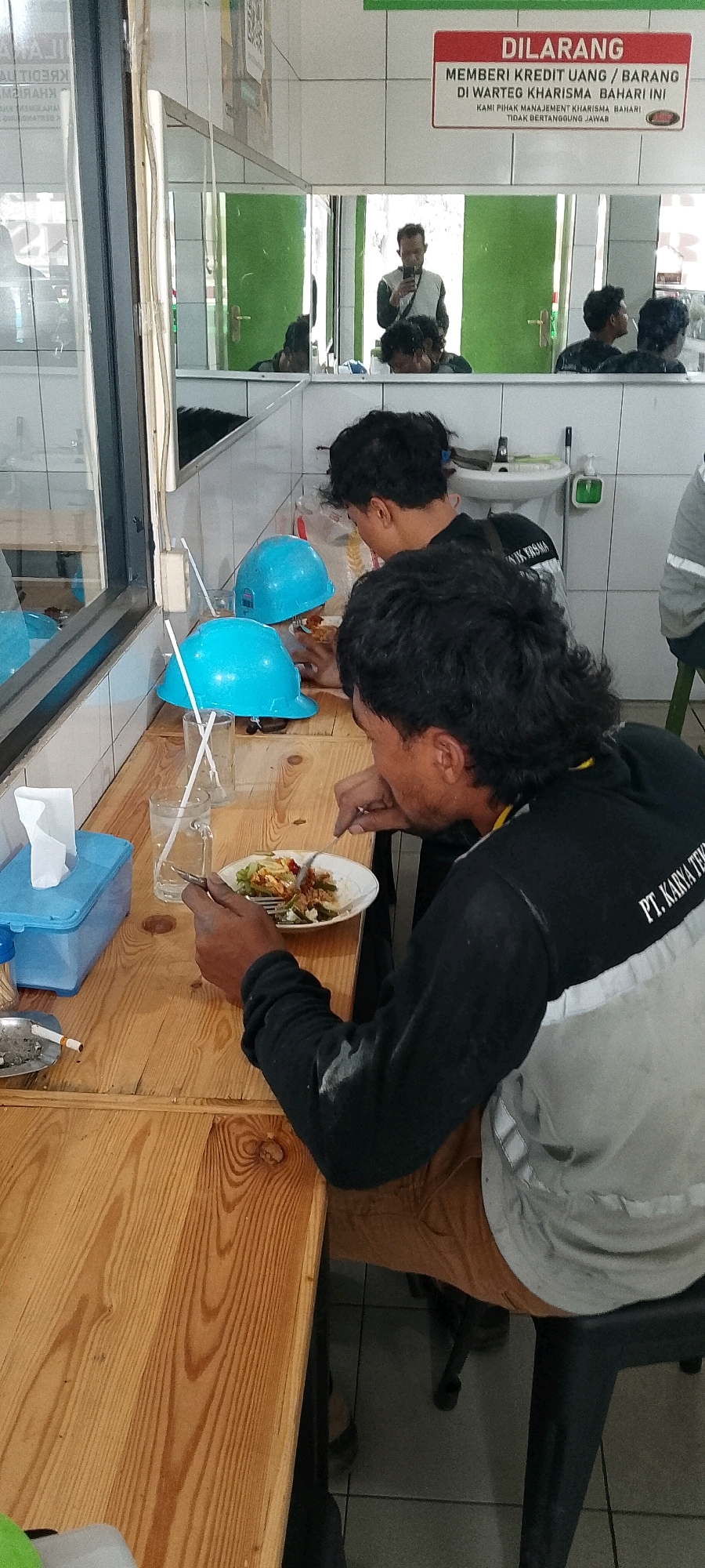 A group of construction workers are eating lunch at a table in a canteen, likely located in Depok, Indonesia. The table is made of light-colored wood with visible grain. Two blue hard hats are placed on the table, along with glasses of water and a tissue dispenser. One worker is actively eating a meal from a white plate, using a fork and knife. Another worker is visible in the foreground, also seated at the table. In the background, a mirror reflects a man in a grey and white shirt taking a picture with his phone, and other people in the canteen. A sign in the mirror reads "DILARANG MEMBERI KREDIT UANG / BARANG DI WARTEG KHARISMA BAHARI INI KAMI PIHAK MANAJEMEN KHARISMA BAHARI TIDAK BERTANGGUNG JAWAB." The overall environment appears to be a simple, functional canteen.