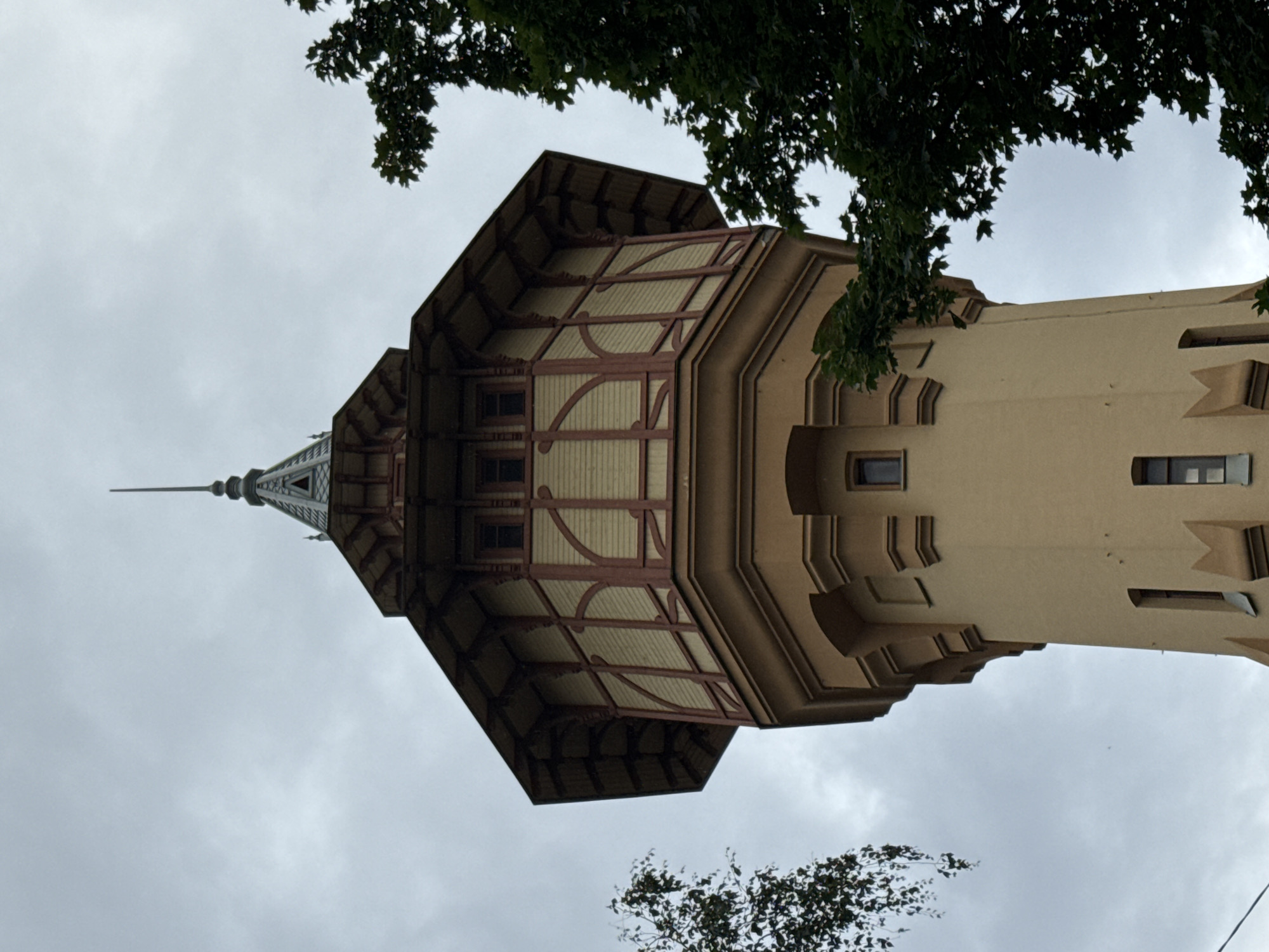 A low-angle, full shot captures the top of a pale orange, octagonal tower against a cloudy, overcast sky. The tower features decorative architectural elements, including a projecting upper section with a light tan exterior and dark red trim. This section has multiple small, dark-paned windows and decorative wooden bracing. The tower is topped with a grey, tiered roof culminating in a thin spire. On the left and right sides of the frame, dark green tree branches with leaves extend into the shot, partially obscuring the tower. The overall scene appears to be an outdoor view of a building, likely a historical landmark or water tower, in an urban setting. The location is identified as Unknown, Russia.