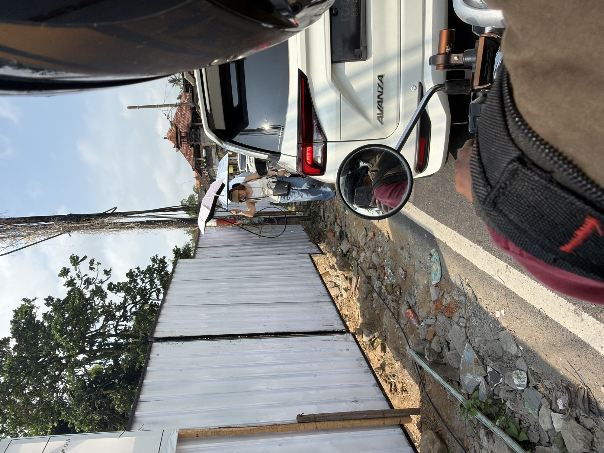 The image shows a street scene in Ubud District, Indonesia, likely near a construction site. The foreground is dominated by the handlebars and part of the seat of a motorcycle, indicating the perspective of the photographer. A white SUV, identified by the "AVANZA" badging on the rear, is parked or stopped in traffic in the middle ground. To the left of the SUV, a woman is walking while holding a light-colored umbrella. She is wearing a white tank top, blue jeans, and a tan baseball cap. Behind her is a white corrugated metal fence, which appears to be part of the construction site. The ground along the fence is littered with construction debris, including rocks and broken bricks, as well as some scattered trash. In the background, traditional Indonesian architecture with orange tiled roofs is visible, along with lush green trees and a bright blue sky with scattered clouds. The overall impression is a snapshot of everyday life on a busy street in a tropical environment.
