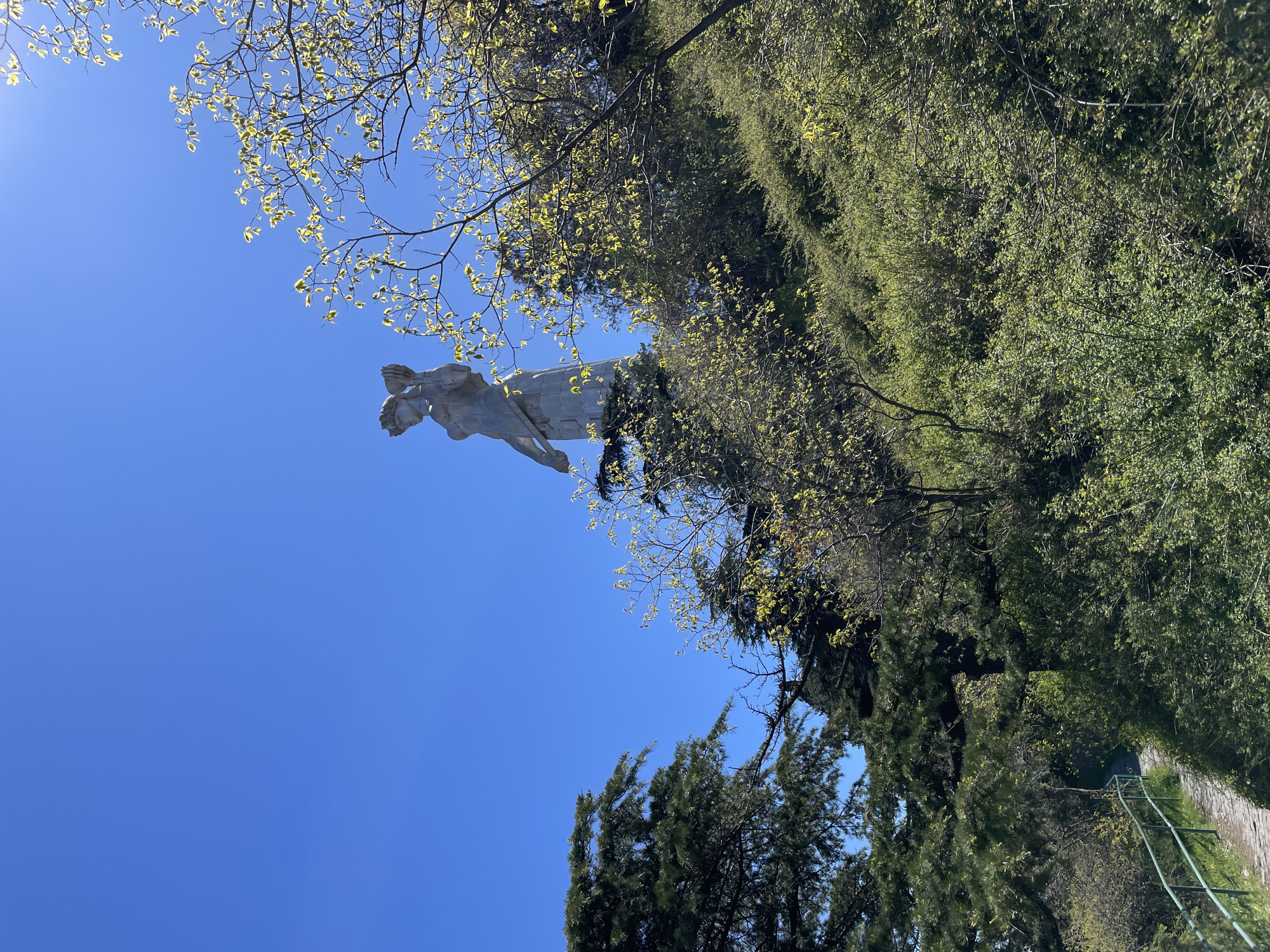 The image shows the Kartlis Deda monument in Tbilisi, Georgia, from a low angle, with the statue set against a clear blue sky. The monument is located on Sololaki hill. Trees frame the view, some with new spring foliage. A pathway with a metal railing is visible in the lower right corner.