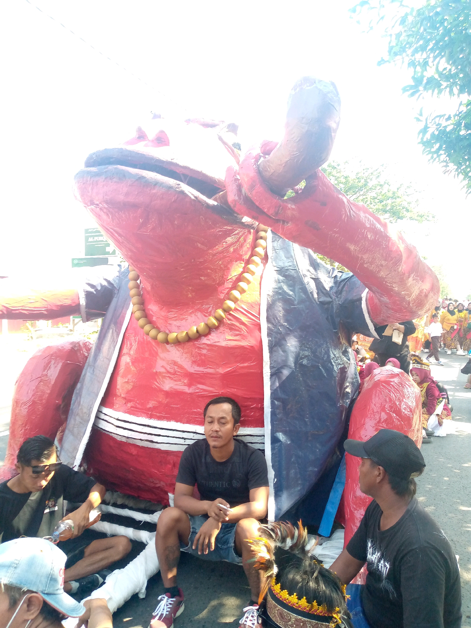 In Sumberwono, Indonesia, a large, colorful papier-mâché effigy of a red, frog-like creature stands prominently in the foreground. The effigy is adorned with a beaded necklace and wears a jacket with blue and dark grey panels. Several individuals are seated around the base of the effigy. Two men in black t-shirts are most visible, one pouring from a plastic bottle and the other holding a cigarette. Another person wearing a black baseball cap is also seated nearby, looking to the right. In the background, a procession of people in traditional attire, including women in yellow and red garments, are visible walking down a street lined with trees. The scene appears to be part of a local festival or parade.