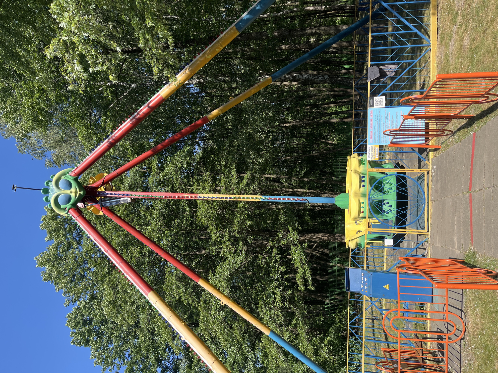The image depicts an amusement park ride featuring a central vertical pole with multiple colorful arms extending outwards. The arms are painted in sections of red, yellow, and blue. At the top of the central pole, a decorative, cartoonish face with large blue and green eyes is visible. The ride is situated outdoors in a park-like setting, surrounded by lush green trees. In the foreground, there are brightly colored fences, predominantly orange and blue with yellow accents, sectioning off the area. A blue control panel with buttons and a yellow warning symbol is visible to the left of the ride's entrance. Signs with text and a QR code are posted on the fence. The ground is a mix of paved pathway and grassy areas. The sky is clear and blue. There are no people visible in the image.