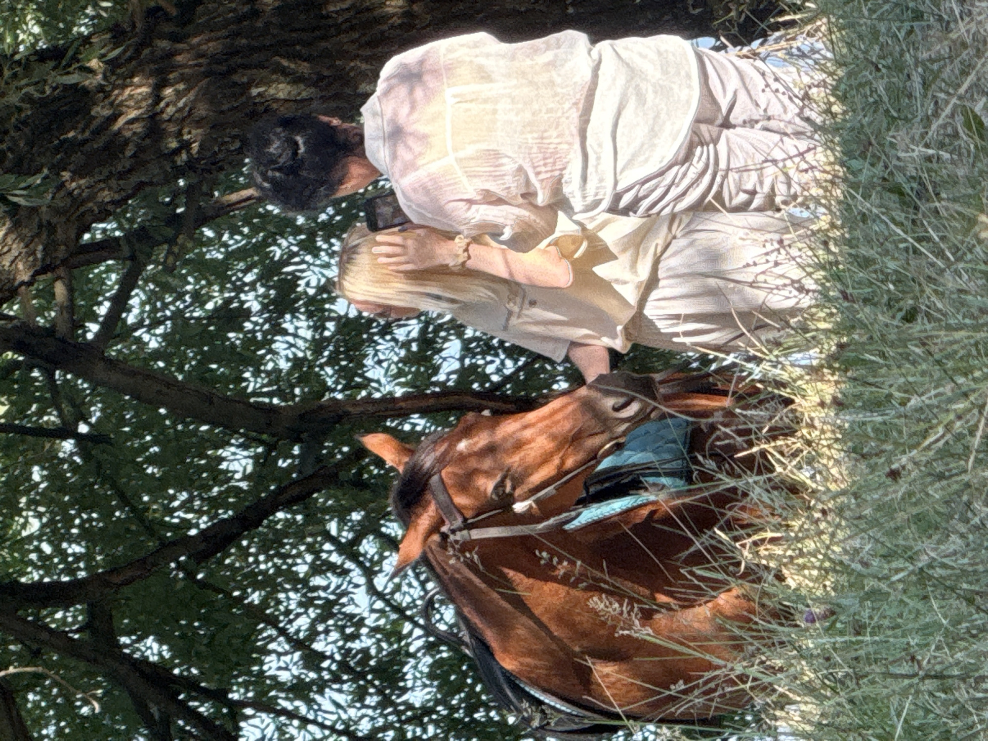 The image shows two individuals standing near a chestnut-colored horse in an outdoor setting. One individual, with blonde hair, is wearing a light-colored shirt and pants. The other individual, with dark hair, is wearing a translucent, light-colored shirt and striped pants. The dark-haired individual appears to be holding a smartphone and taking a photo or video of the blonde-haired individual. The horse is wearing a bridle and a saddle with a teal-colored saddle pad. The environment suggests a natural landscape with trees and tall grass. The scene is bathed in sunlight, with dappled light filtering through the tree leaves. The location is Zaprudnoe, Ukraine.