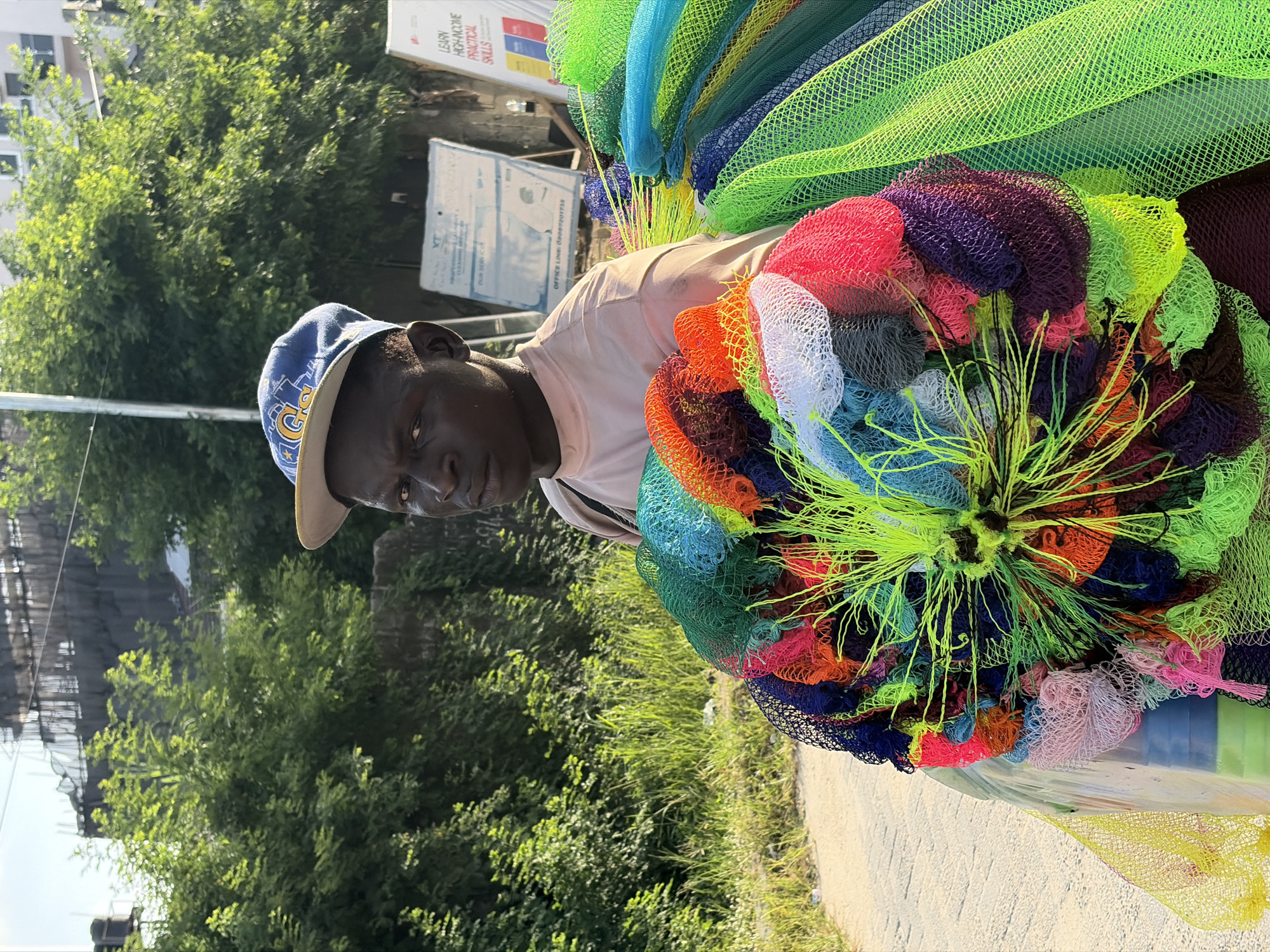 A man wearing a blue baseball cap and light pink shirt stands on the side of a road in Igboefon, Nigeria. He carries a large assortment of colorful netted sponges. The sponges are arranged in a way that some are on top of his head and shoulders, while others are bundled and held in front of him. The sponges have a variety of colors, including shades of green, blue, red, orange, purple, and white. The background includes lush green vegetation and a partially visible sign with text. The road is paved and there's a grassy area beside it. The lighting suggests it is daytime.