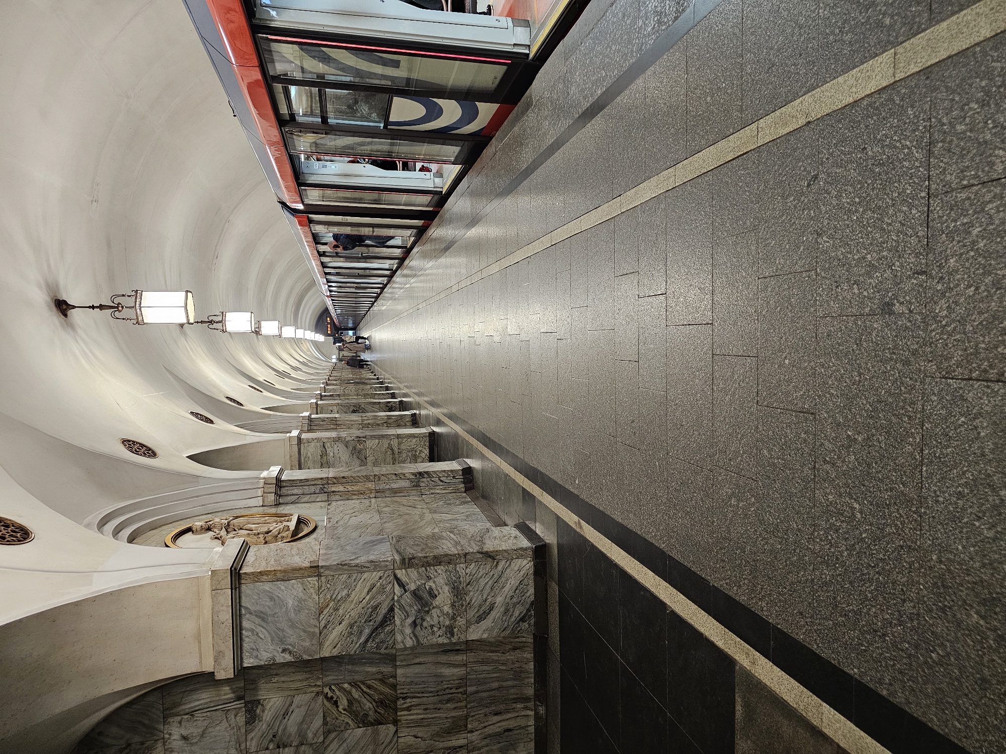The image depicts a long, narrow view of a Moscow Metro station platform in Russia. The platform features a grey tiled floor with a stripe near the edge. On the left side, a curved, white ceiling with recessed lighting fixtures stretches into the distance. Below the ceiling are marble walls with raised platforms and decorative elements. A red and grey metro train is visible on the right. The vanishing point of the perspective is far down the platform, where people are visible in the distance.