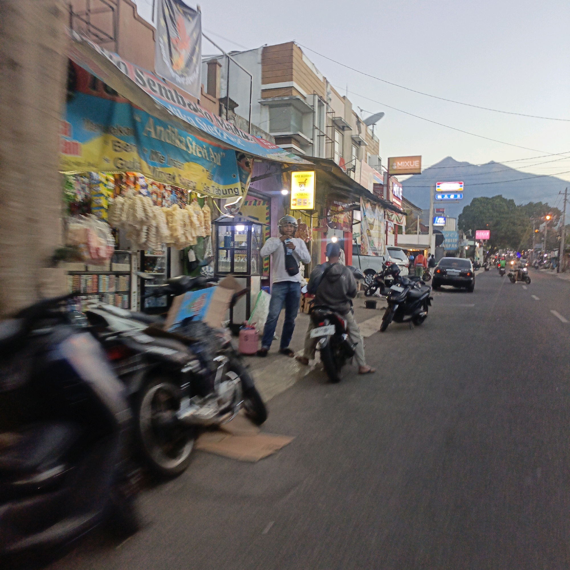 The image depicts a street scene in Salatiga, Indonesia, likely in the late afternoon or early evening, given the lighting. The foreground is dominated by a blurred motorcycle on the left. The midground features a street-level shop with awnings and various goods displayed, including packaged snacks and what appear to be dried food items. Two men are visible near the shop: one wearing a white long-sleeved shirt, jeans, and a helmet, holding a blue bottle, stands near the shop entrance. Another man, wearing a grey hoodie and pants, is seated on a motorcycle to the right of the first man. Further down the street, other motorcycles and a car are visible, suggesting moderate traffic. Buildings line the street, and a distant mountain range is visible against the sky. Signs for "MIXUE" and other businesses are present on the buildings. The overall atmosphere is that of a busy commercial street in a city with a mountainous backdrop.