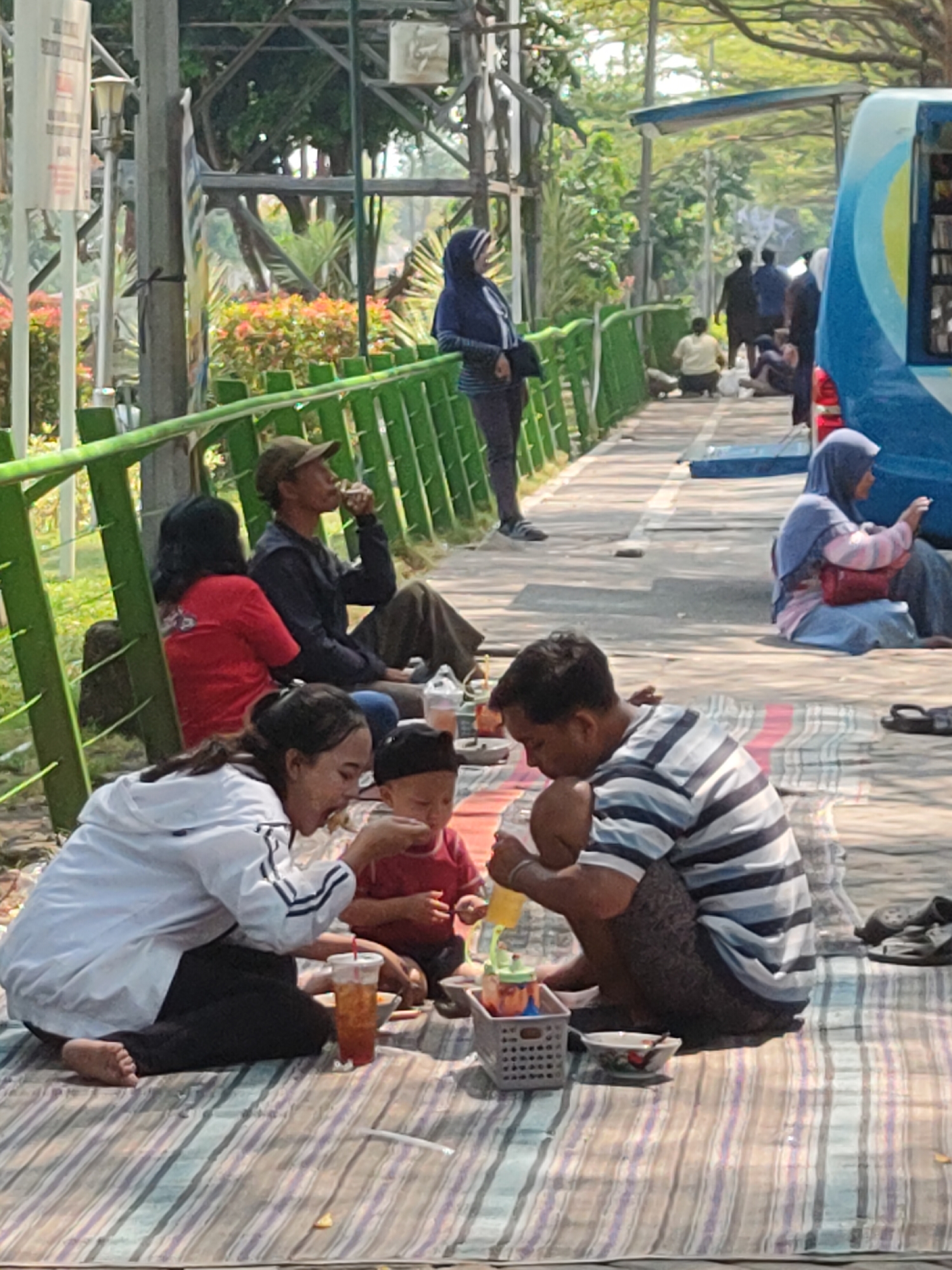 In Ngasem, Indonesia, a family is having a picnic on a striped mat outdoors. A woman in a white jacket and black pants is feeding a young child in a red shirt, who is wearing a black cap. Next to them, a man in a striped t-shirt and patterned shorts is holding a yellow drink. Behind them, a man in a brown hat and dark shirt is seated, and a woman in a red shirt is sitting behind him. In the background, several other people are walking and sitting, and a blue and yellow bus is parked. The environment is bright and sunny, with trees and greenery visible.