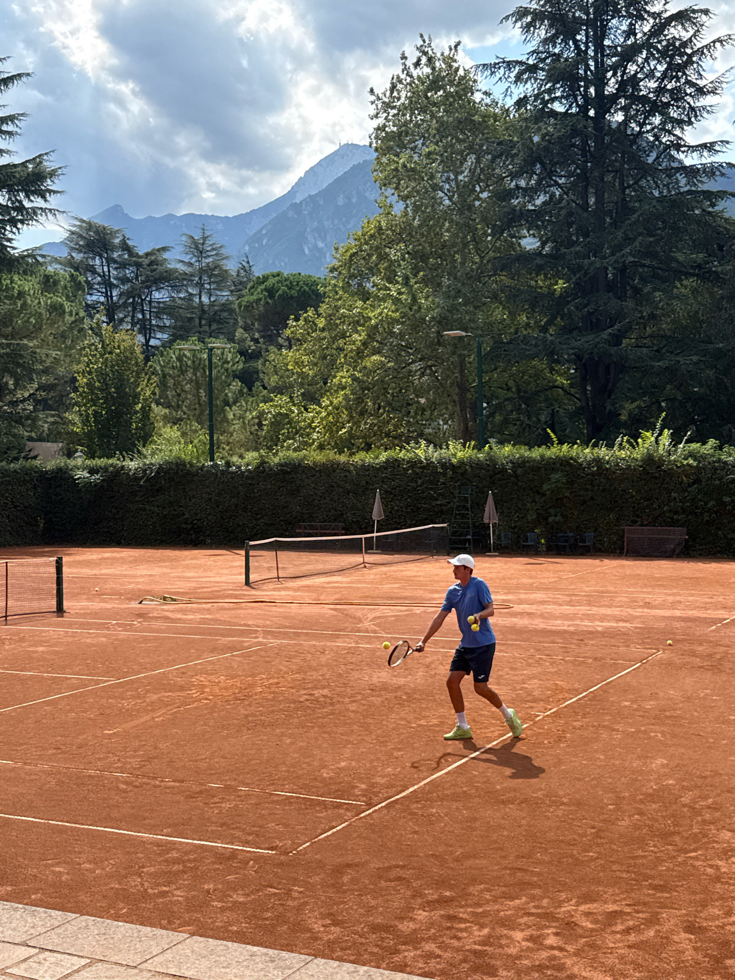 A young male tennis player in a blue shirt, dark shorts, a white cap, and neon green tennis shoes is in motion on an outdoor clay tennis court, holding a tennis racket and three tennis balls. He is right-handed and appears to be in the process of serving or hitting a ball. Two tennis balls are in the air near him, and one is on the ground. The court is surrounded by a hedge and trees, with mountains visible in the background under a partly cloudy sky. The scene is set in Trento, Italy, likely at a tennis club.