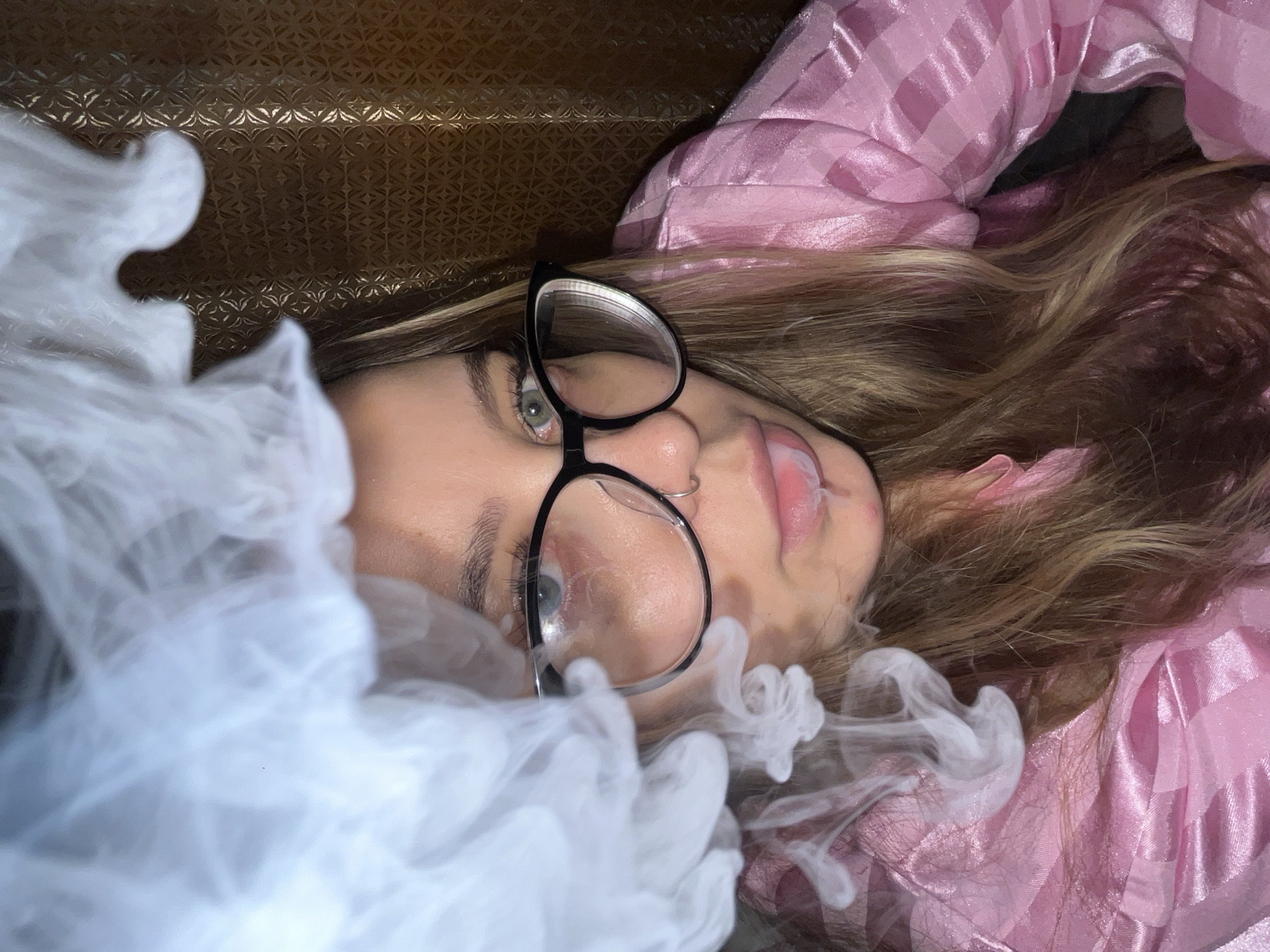 The image shows a young woman lying on a patterned surface, wearing glasses, and blowing smoke. She is wearing a pink checkered garment. The environment is dark. The location is unspecified; the reference to Novopokrovskaya, Russia, is not evident in the image.