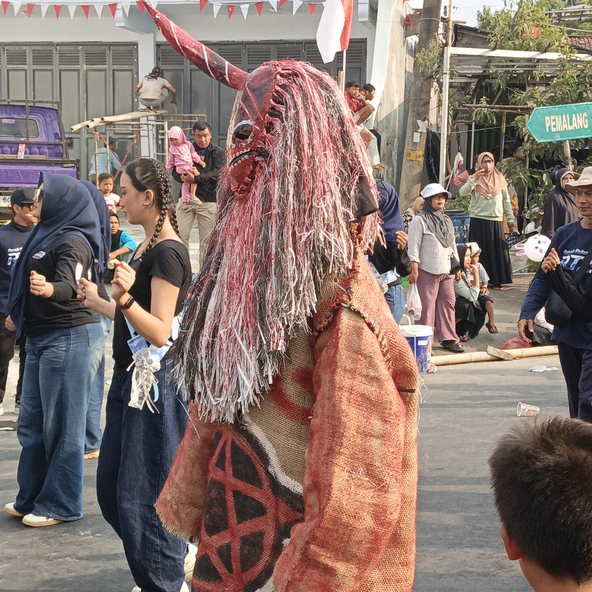 In Randudongkal, Indonesia, a street festival is underway. The central focus is a person in a striking costume, wearing a large, red, demonic mask with long, stringy hair. The costume appears to be made of burlap and fabric, with a red and black geometric pattern on the chest.
Surrounding this figure are numerous people of various ages. To the left, two young women are visible, one wearing a dark blue hijab and a black t-shirt, the other in a black t-shirt with braided hair. Behind them, a purple truck and scaffolding are present. To the right, a green directional sign with the text "PEMALANG" is visible. Other individuals in the background are dressed in casual clothing, some holding children. The scene is set on a paved street with buildings and trees in the background, suggesting an urban or semi-urban environment. The atmosphere appears festive, with some attendees observing the masked figure. Red and white triangular flags are strung across the top of the frame, indicating a celebration.