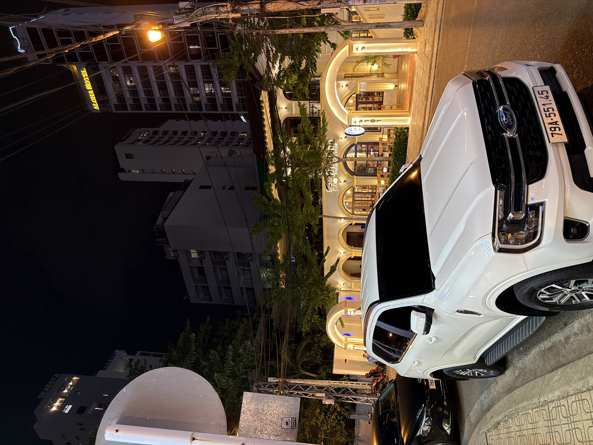 The image captures a nighttime scene on a city street in Nha Trang, Vietnam. In the foreground, a white Ford SUV is parked on the right side of the road, its headlights and grille illuminated. To the left of the SUV, a black car is also parked. Behind the vehicles, a building with arched entrances and illuminated windows stands out. The building appears to be a restaurant or establishment, with outdoor seating visible through the windows and arches. The facade is decorated with warm, glowing lights. Tall buildings, one of which has "ALOHA HOTEL" illuminated in yellow lettering, are visible in the background, indicating an urban environment. The sky is dark, suggesting it is nighttime. Electrical wires are visible crisscrossing the scene, typical of urban streetscapes. There are a few people visible in the background, near the entrance of the illuminated building. The street is paved, and a sidewalk with decorative patterns is present. The overall atmosphere is that of a lively commercial district at night.