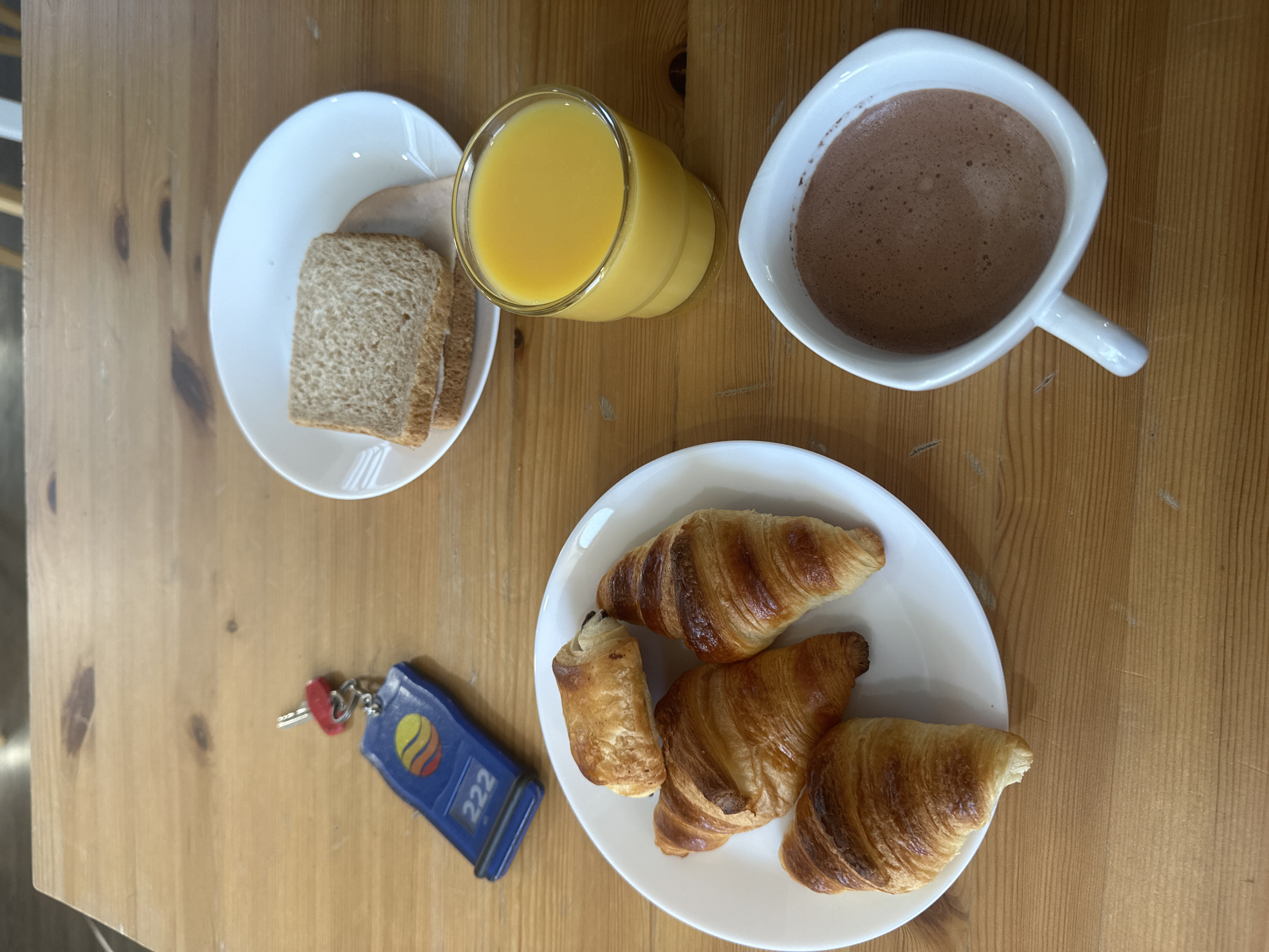 On a light-colored wooden table, a breakfast arrangement is set. A white plate holds two slices of wheat bread and what appears to be a thin slice of meat. A glass of orange juice sits beside the plate. A white, angular mug filled with hot chocolate is placed near the juice. A separate white plate contains four pastries: three croissants and one chocolate-filled pastry. A blue key fob with the number "222" printed on it rests next to the plate of pastries.
While the image suggests a breakfast setting, the exact location in Ramonville-Saint-Agne, France, cannot be confirmed.