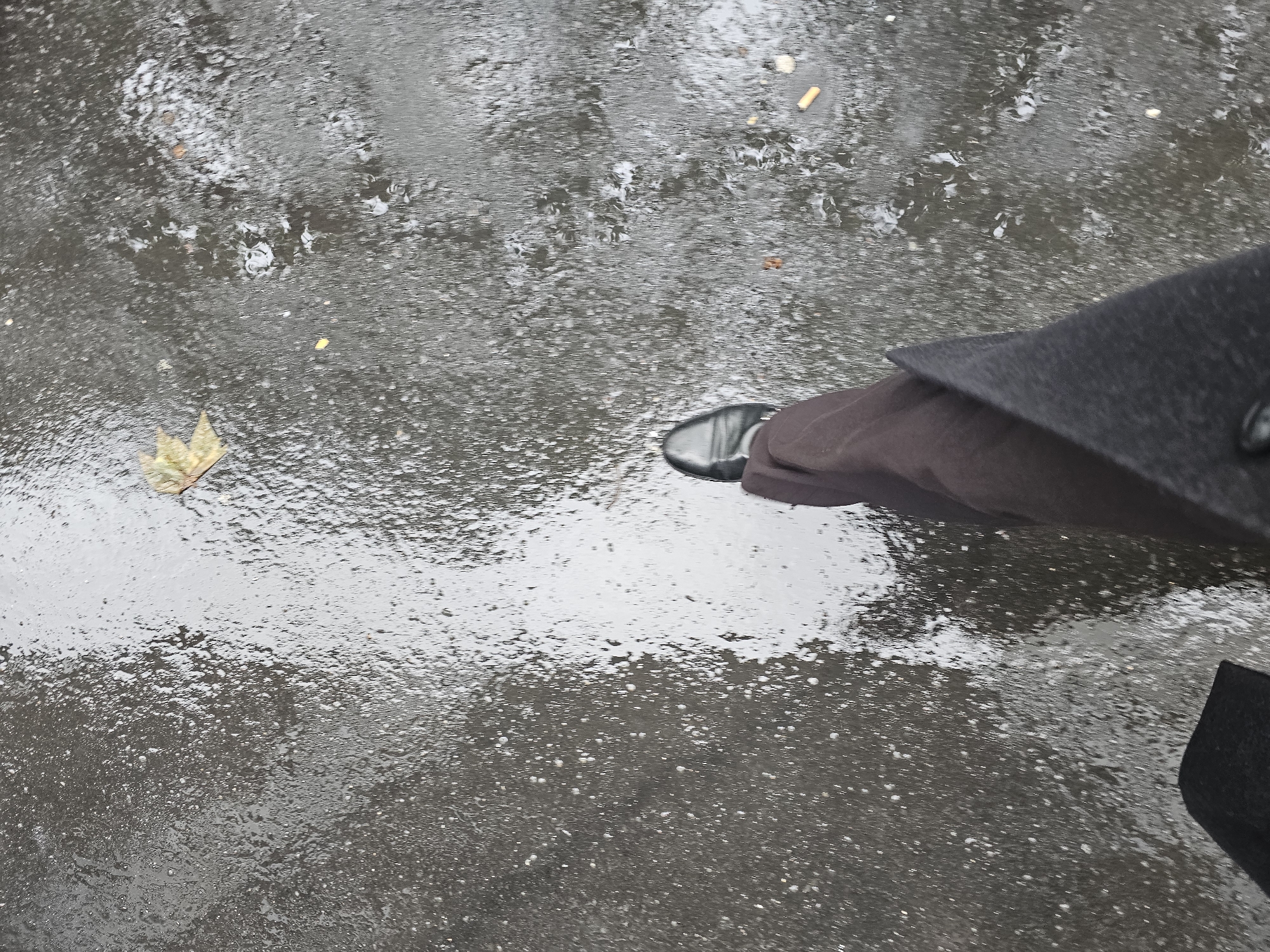 A top-down shot reveals a rain-slicked Parisian street. Predominantly grey, the wet asphalt reflects light, interspersed with debris like a fallen leaf and cigarette butts. A person's lower body is visible, dressed in brown trousers, a dark coat, and a black leather shoe. The coat's button is partially visible. The scene suggests a wet, overcast day in Paris.