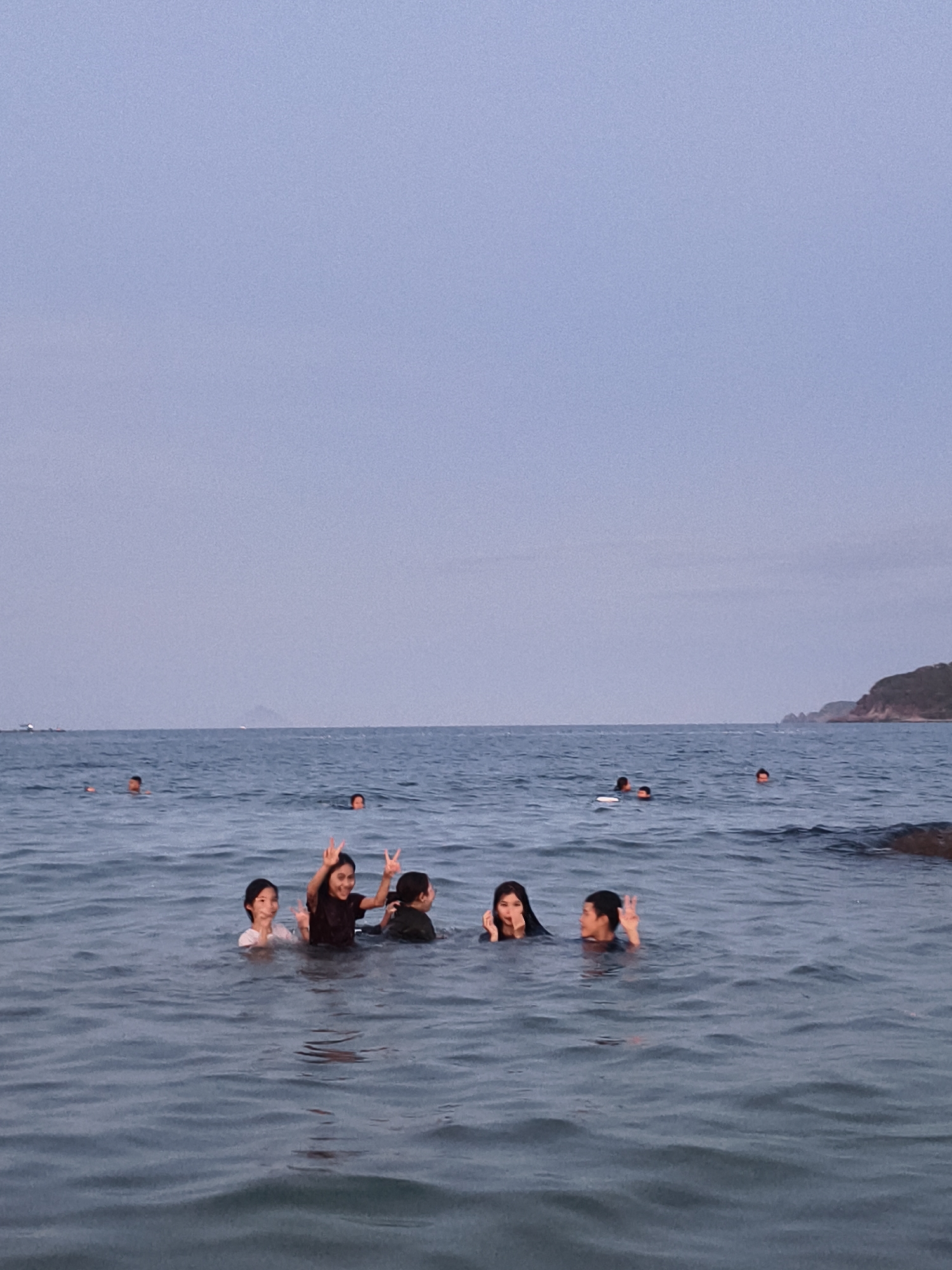 In Vĩnh Lương, Vietnam, a group of individuals are swimming in the sea under an overcast sky. Several people are visible in the water, with most of their bodies submerged. In the foreground, three young women are partially submerged, with one on the left wearing a white top, the middle one in a dark top, and the one on the right in a dark top. The middle woman is making a peace sign with her right hand. To the right of the three women, a young person in a dark top is also swimming, facing towards the camera and making a peace sign with their left hand. In the middle ground, several other individuals are scattered in the water, with only their heads visible. In the background, a distant headland covered in vegetation is visible on the right, and a faint mountainous silhouette is seen on the horizon to the left. The sea is calm with gentle waves.