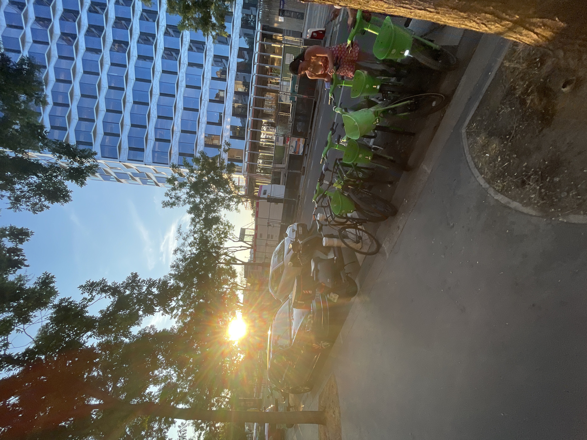 The image captures a sun-drenched street scene in Paris, France. In the foreground, a row of bright green rental bicycles is parked along the sidewalk. A woman in a patterned dress is interacting with one of these bicycles. Further back, several parked cars and a scooter are visible on the street. The background features a modern, glass-fronted building with a distinctive geometric facade. Trees line the street, with the setting sun casting long shadows and creating a lens flare effect. The overall environment suggests an urban setting during golden hour.