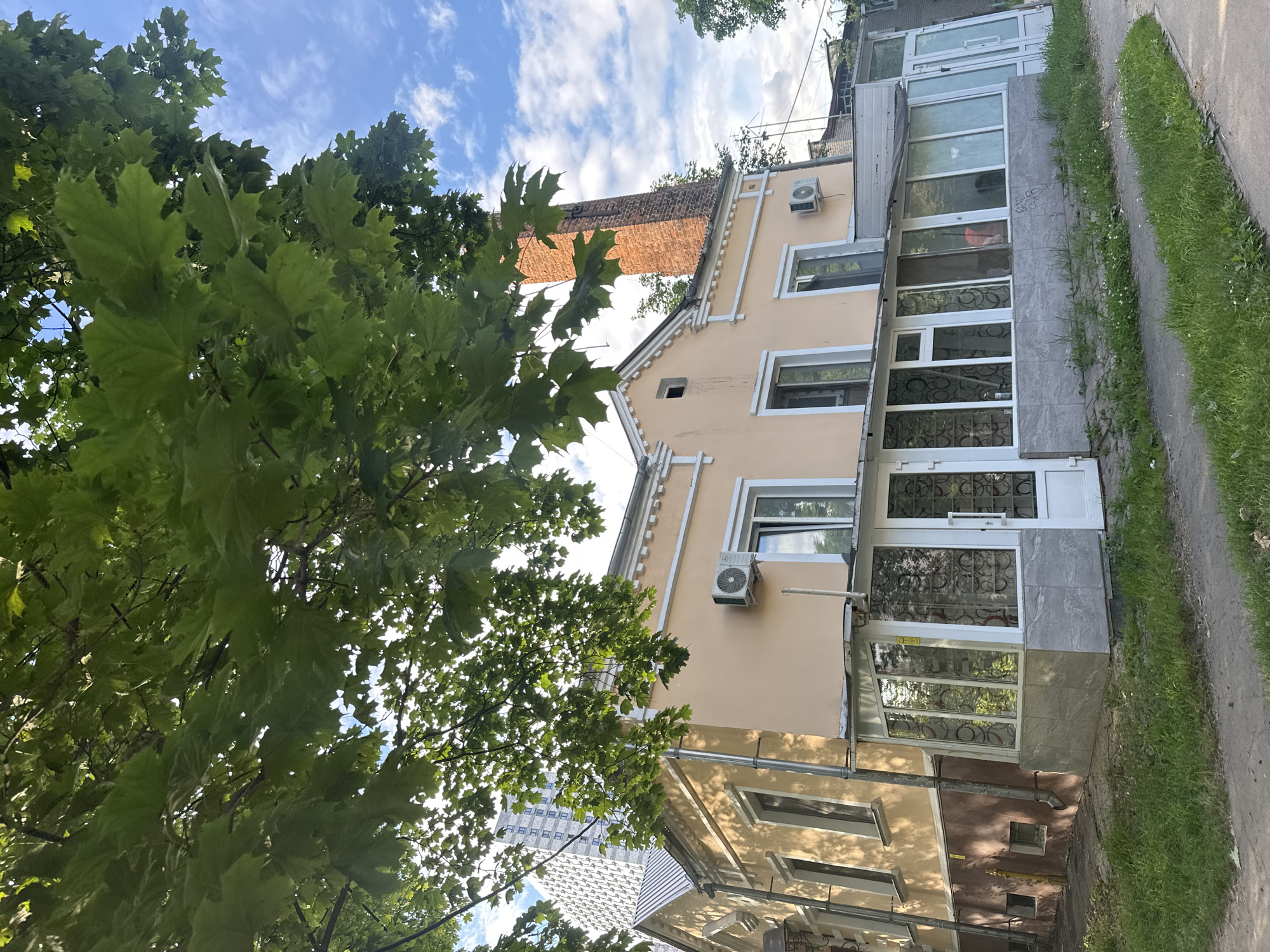 This is a vertical, eye-level, outdoor shot of a peach-colored building with a tiled roof and windows on multiple floors. The building is partially obscured by the branches and green leaves of a large tree in the foreground. The sky is visible behind the tree and building, with white clouds against a blue background. A brick chimney is visible on the roof of the building. The building appears to be a residential or commercial space, with large windows and glass doors at street level. A sidewalk runs along the side of the building, with green grass and some small white flowers growing along its edge. The lighting suggests it is daytime, possibly late afternoon due to the shadows. The overall scene is serene and suburban. No people or specific activities are depicted. No text is visible in the image. The location context is Kotelniki, Russia.