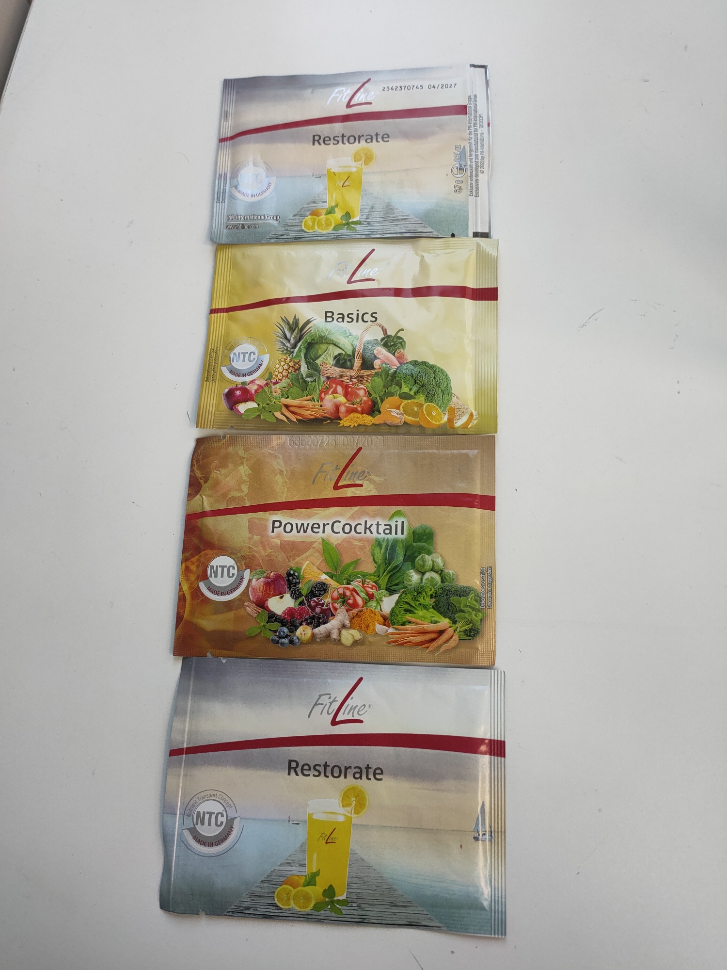 The image displays four individual sachets of "FitLine" nutritional supplements, arranged vertically on a plain white surface. From top to bottom, the products are identified as "Restorate," "Basics," "PowerCocktail," and a second "Restorate" sachet.
The two "Restorate" sachets are silver-grey, each featuring a visual of a glass of yellow beverage with lemon slices on a wooden pier, set against a tranquil water backdrop. The top "Restorate" sachet shows legible text including an expiration date of "04/2027," "6.7 g 0.24 oz," and "Exclusively developed and manufactured by PM-International Group."
The "Basics" sachet is yellow and illustrates a generous assortment of fresh produce, such as pineapple, cabbage, broccoli, apples, carrots, and oranges, presented in a basket.
The "PowerCocktail" sachet has a bronze-brown color and depicts a rich variety of fruits, vegetables, and spices, including berries, apples, spinach, broccoli, carrots, ginger, and turmeric.
Each sachet prominently displays the "FitLine" brand logo and a circular "NTC" (Nutrient Transport Concept) seal, emphasizing "MADE IN GERMANY," indicating the product's origin and proprietary concept. Other visible text includes "Dekorationsvorschlag" (Decoration suggestion) on the top sachet.
The scene is brightly lit, suggesting an indoor setting like a table or counter, presenting the products clearly. No people are present, and no activity is occurring beyond the static display of the supplement packets. The products are designed and manufactured by PM-International Group, with the general location context being Sochi, Russia.
