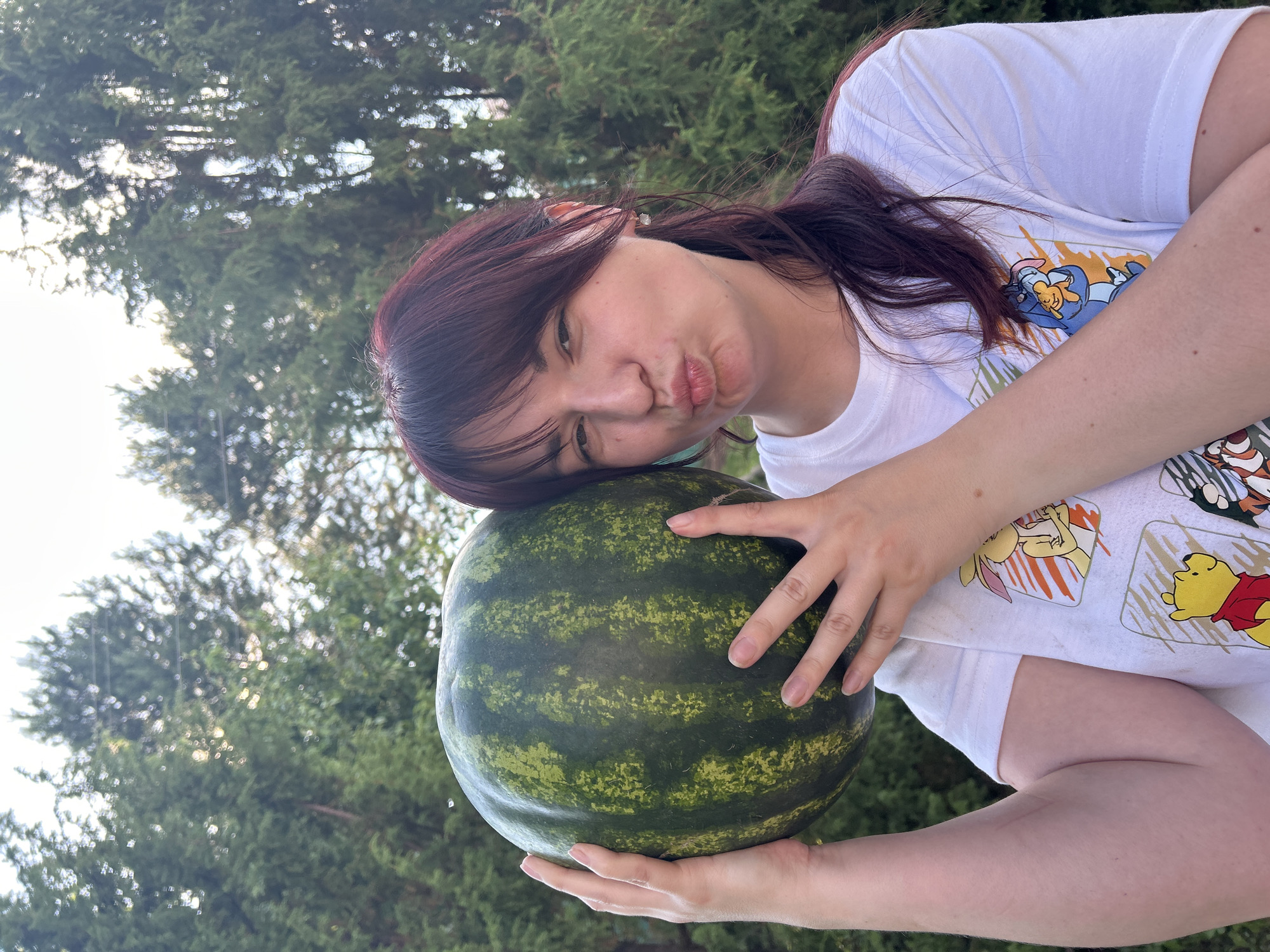 A young woman with dark red hair and bangs is holding a large watermelon in front of her face, puckering her lips as if for a kiss. She is wearing a white t-shirt with cartoon characters printed on it. The background is a blurry image of evergreen trees, suggesting an outdoor setting. It is not possible to determine the location in the city of Fokino, Russia from the image.