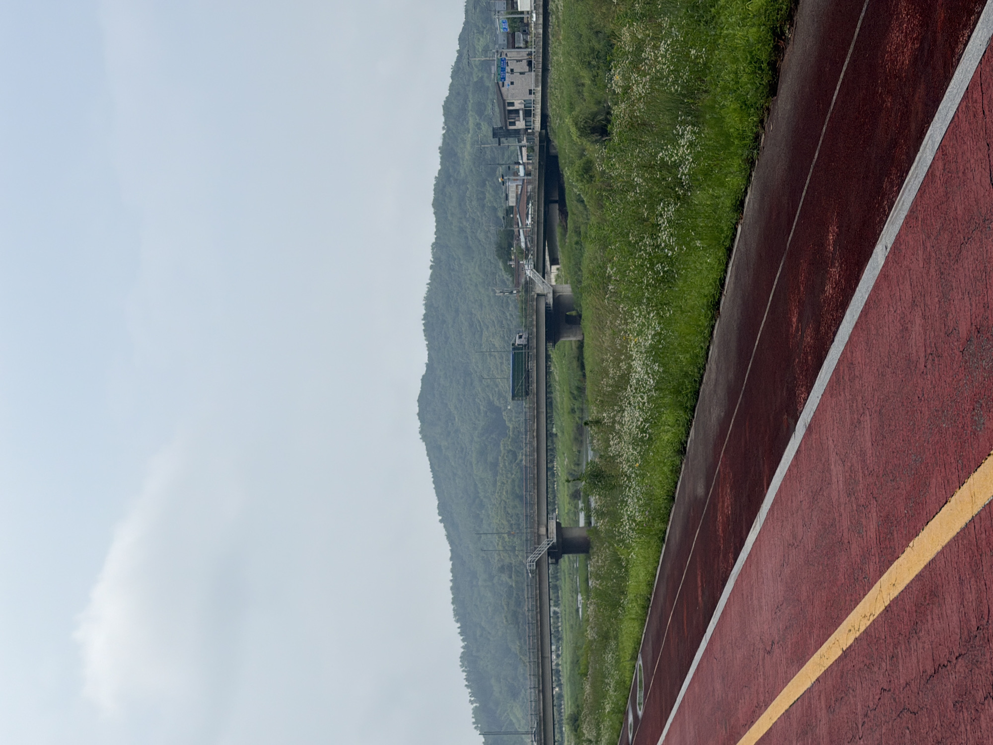 The image captures a scene in Dongducheon-si, South Korea, featuring a running track in the foreground. The track is a deep red color with a distinct yellow line running along its edge, and a white line further out. Beyond the track, a lush green field is visible, dotted with numerous small white wildflowers. In the midground, a river or stream flows, spanned by a concrete bridge. A truck is visible on the bridge, moving from left to right. Further in the background, a verdant, tree-covered hill rises, with buildings and more infrastructure visible at its base and along the road on the bridge. The sky is overcast and hazy, with a pale, muted light.