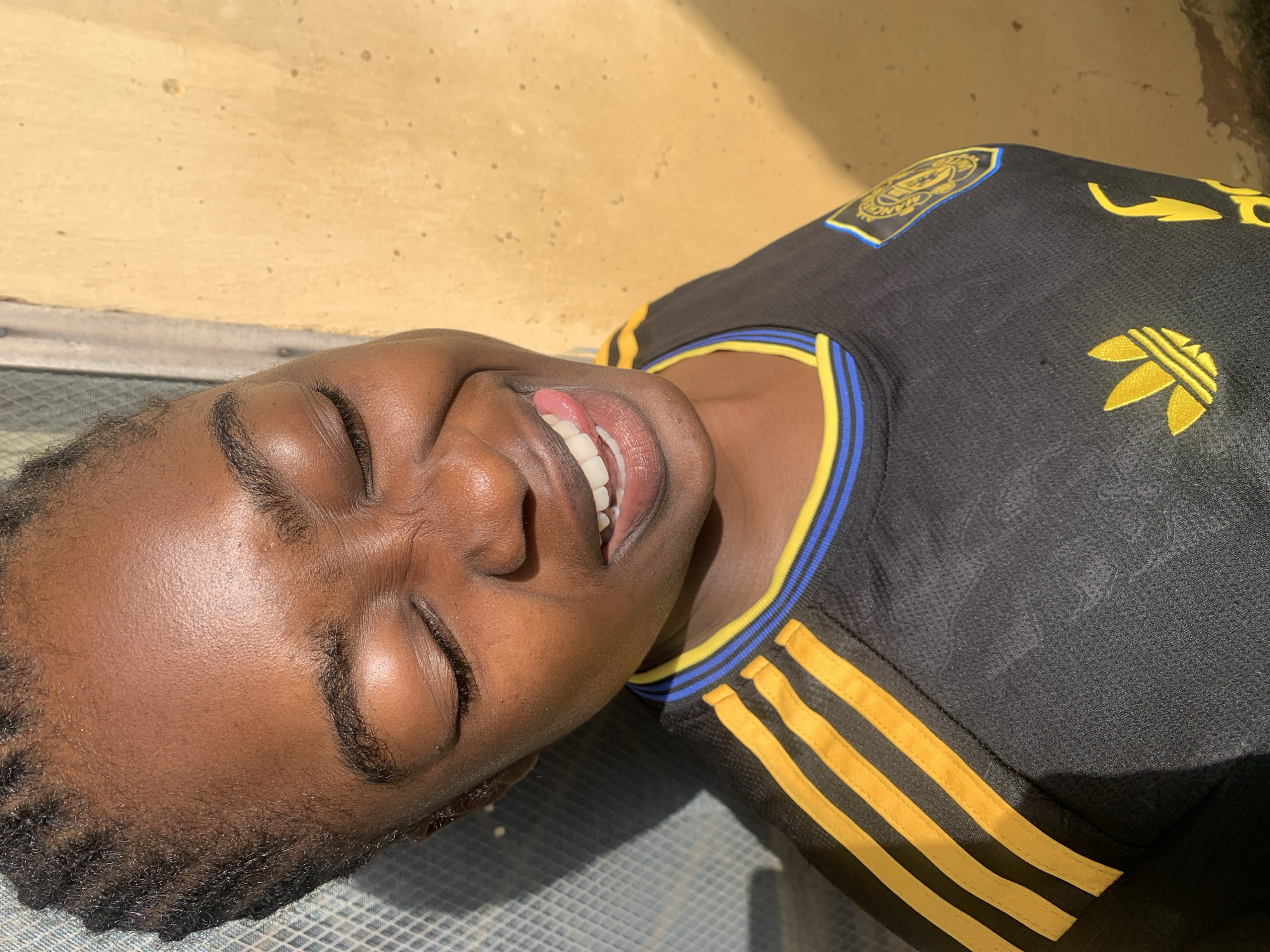 A young Black female with braided hair is shown in a close-up shot, wearing a black jersey with yellow and blue trim. Her eyes are closed, and she is sticking her tongue out playfully. The background consists of a yellow, textured wall and a blue, grid-patterned surface. The lighting suggests it is daytime, with a shadow falling on the left side of the image. This photograph was taken in Rafin Par, Nigeria.