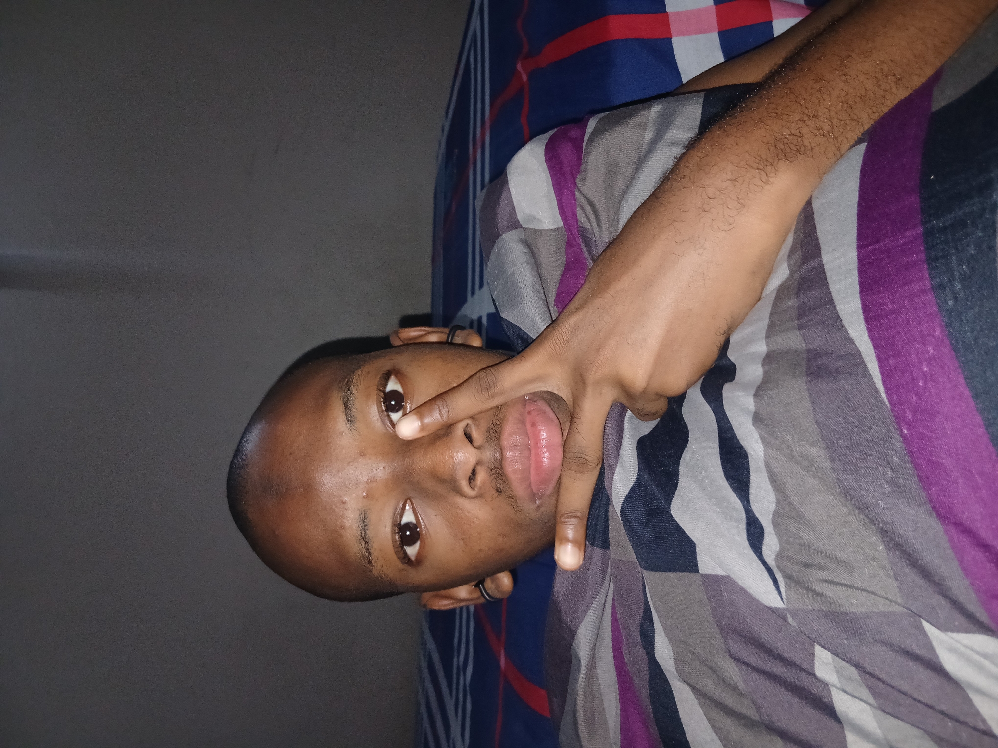 A medium shot captures a young Black male lying down, possibly on a bed covered with a striped duvet. His head is tilted slightly to his left, and he is looking directly at the camera. He has a shaved head and is wearing a black hoop earring in his left ear. His right hand is raised, with two fingers extended in a peace sign, touching his cheek. His lips are slightly parted. The background is a plain, dark wall. The duvet has a geometric pattern with stripes in shades of blue, purple, grey, and white. There are no identifiable landmarks or cues to suggest a specific location in Lagos, Nigeria.