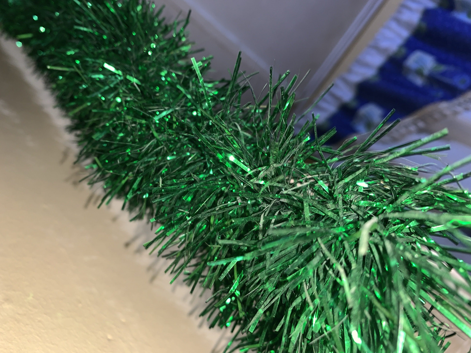 A close-up, low-angle shot shows a strand of bright green tinsel garland. The garland is draped diagonally across the frame, with the closest portion in sharp focus and the background softly blurred. The tinsel has a shimmering, metallic quality, with individual strands reflecting light. The background consists of a beige-colored wall and a glimpse of a blue fabric with white floral patterns. The scene appears to be indoors, possibly in a residential setting. There is no indication of the specific location within Rafin Par, Nigeria.