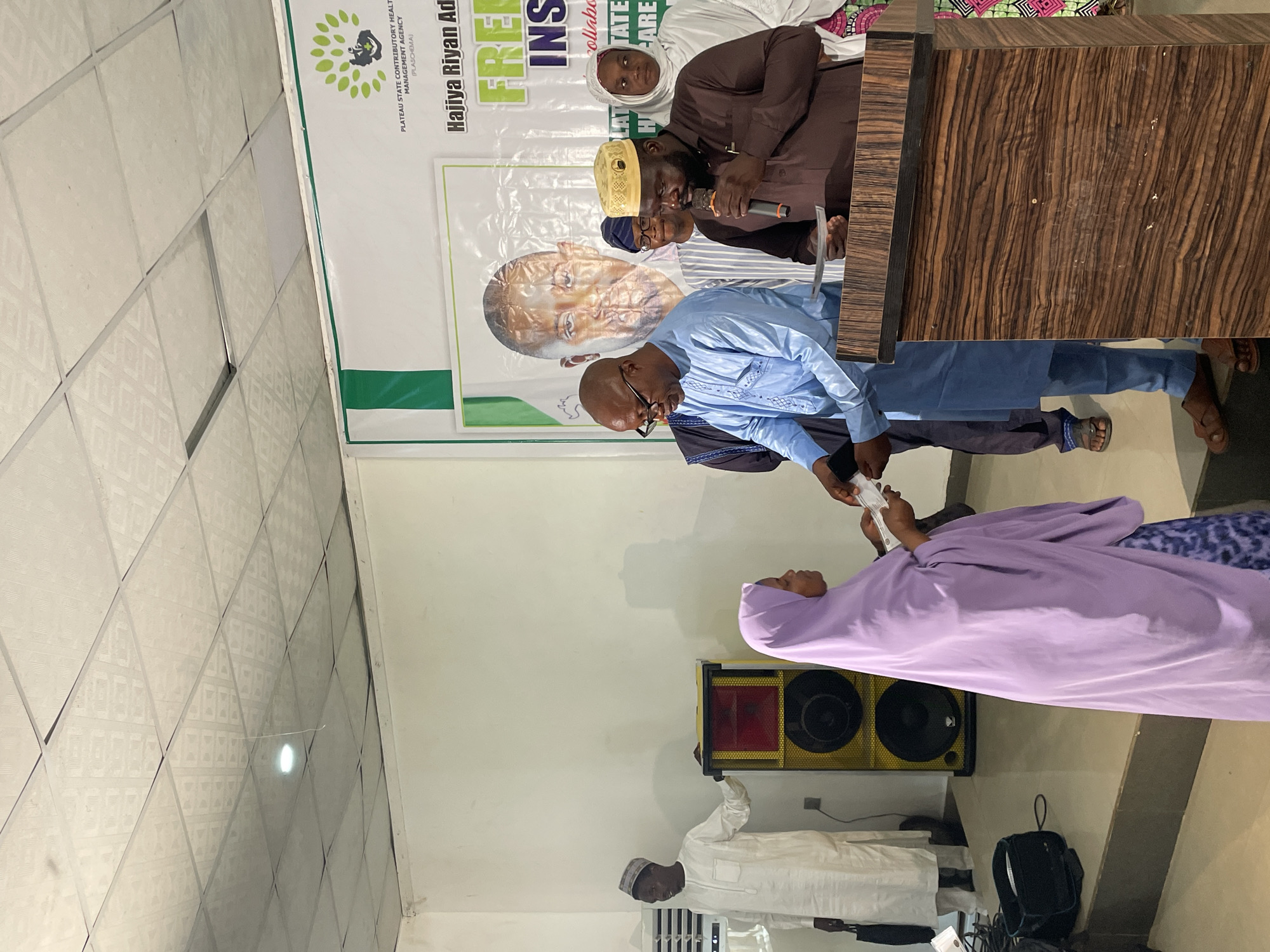 A medium shot shows a room during a public event in Jos, Nigeria. A man wearing a light blue shirt and pants is handing a wad of cash to a woman in a purple headscarf and gown, while another man wearing a brown robe speaks into a microphone while standing behind a wooden podium. Behind them is a sign bearing the words "Hajiya Riyan Ad," which indicates that this is an event sponsored by the Hajiya Riyan Adult and Maternal Healthcare Agency. A banner is also behind them with the green and white colors of Nigeria. A black speaker box is visible next to the woman in the purple gown. The people present seem to be dressed in traditional Nigerian clothing.